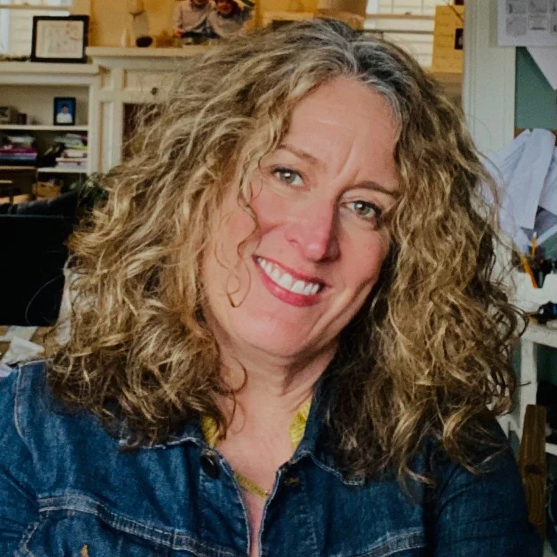 A woman with curly blonde hair smiles at the camera, wearing a denim jacket in an indoor setting with shelves and papers in the background.