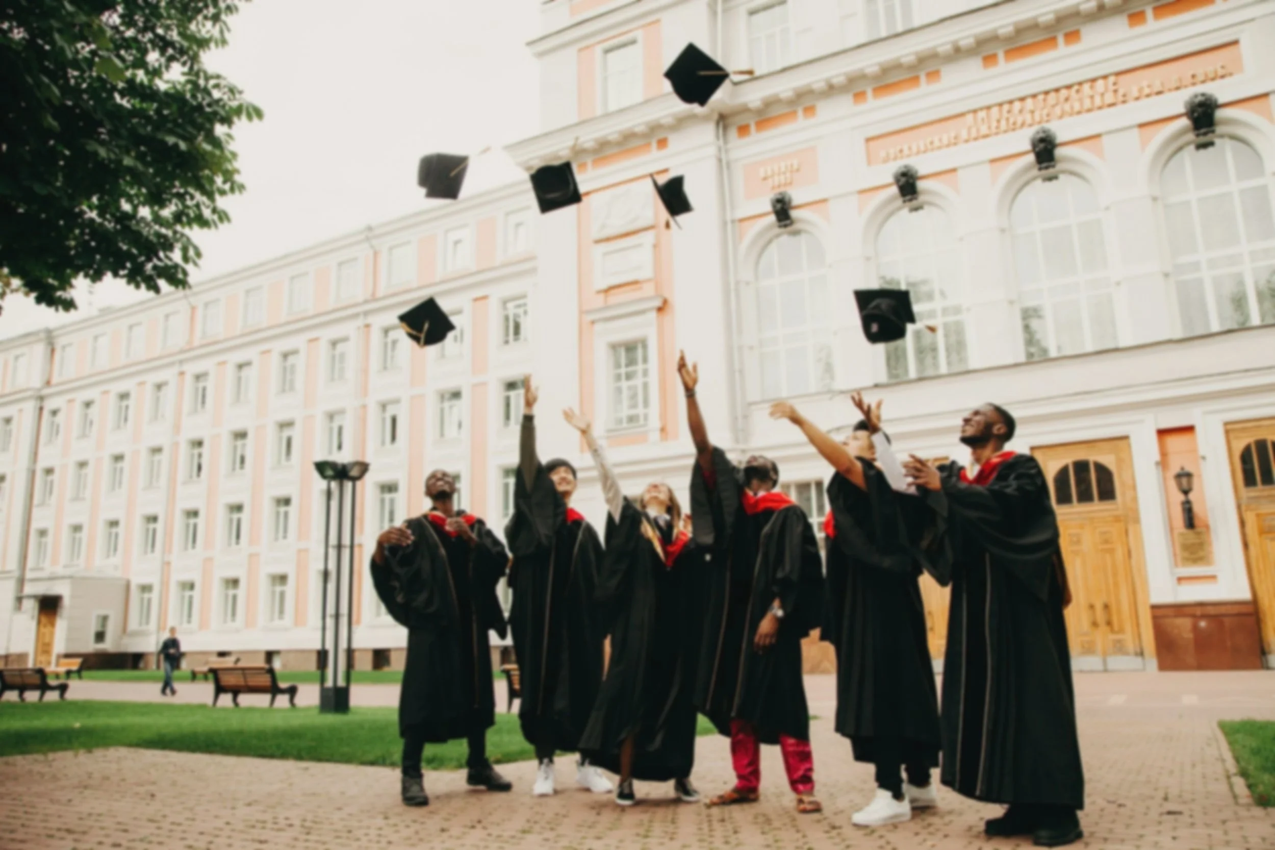 Group of graduates in caps and gowns celebrating outdoors in front of a pastel-colored building.