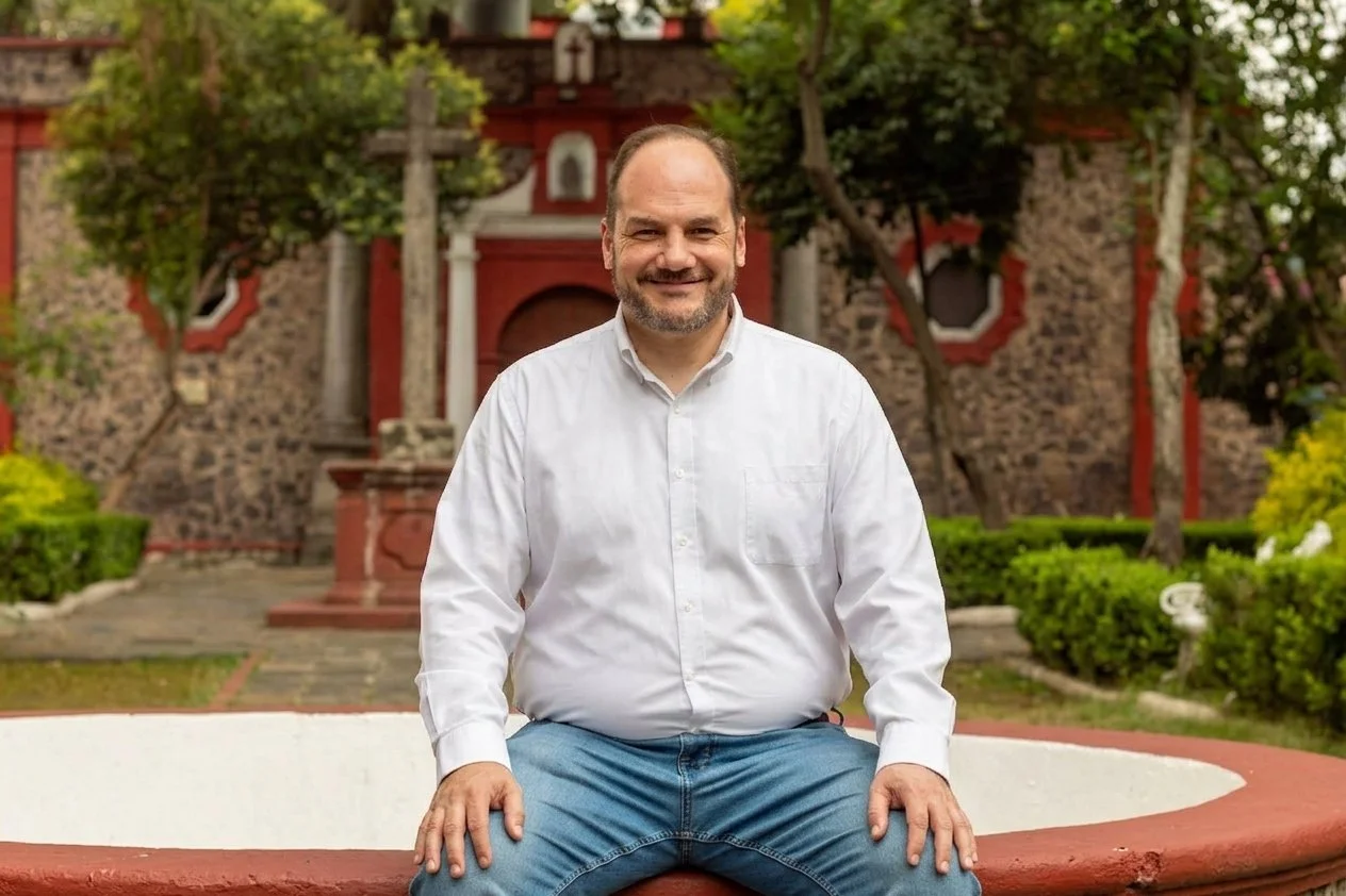 Un hombre sentado en un borde circular en un jardín con árboles y un edificio de piedra de fondo, sonriente y con camiseta blanca y jeans azules.