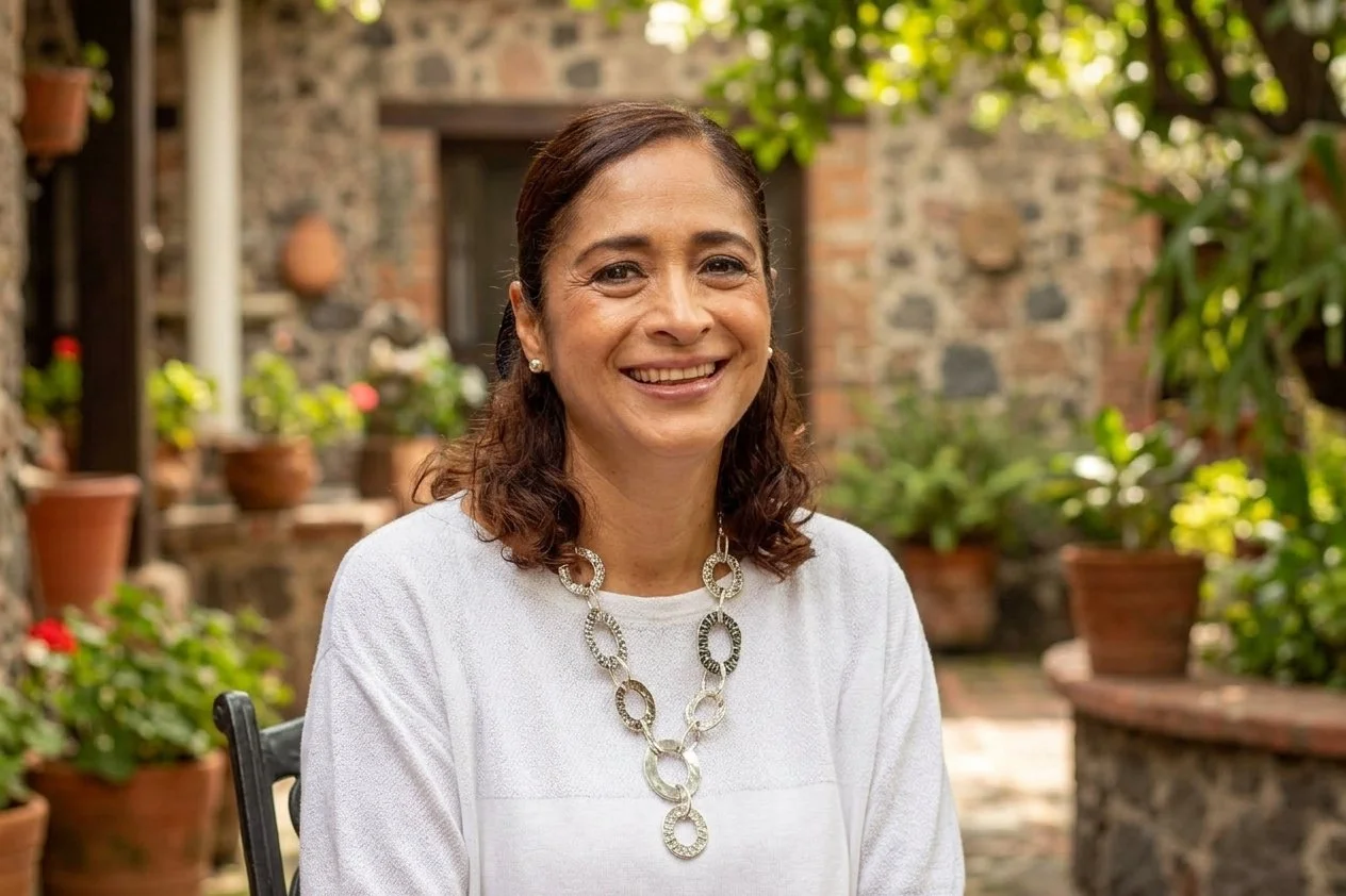 Retrato de una mujer sonriente en un jardín con plantas en tiestos de barro y paredes de piedra.