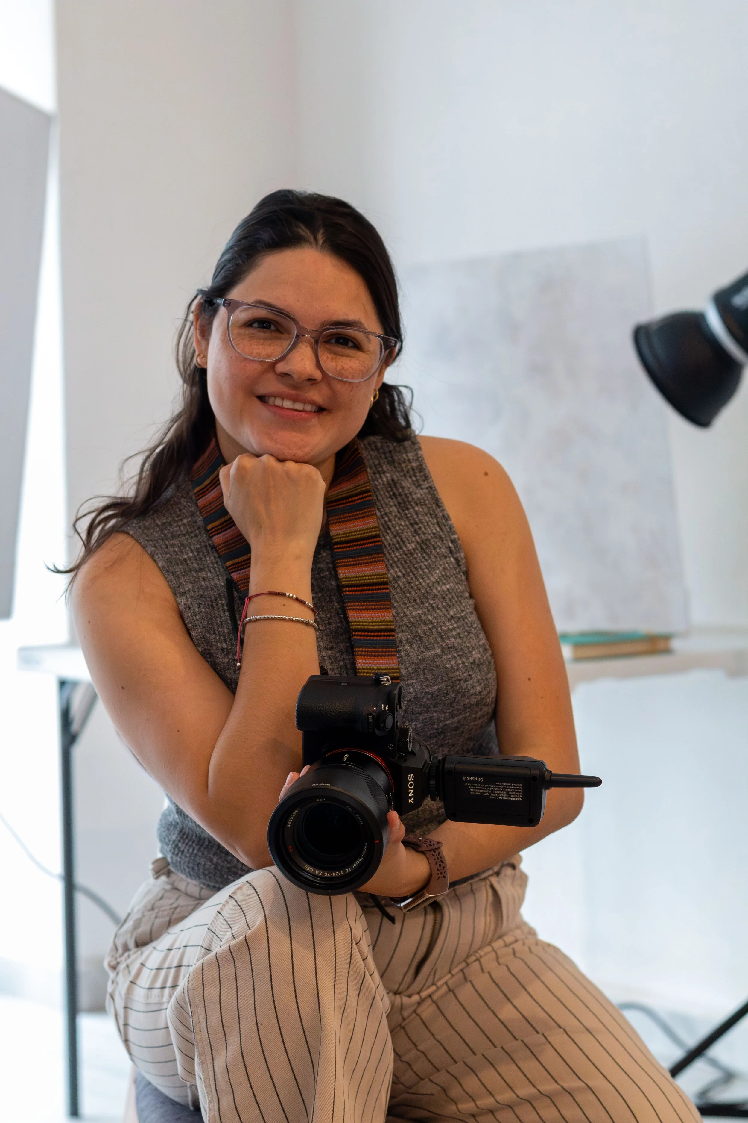 Mujer con gafas, sosteniendo una cámara en un estudio fotográfico, sonriendo a la cámara.