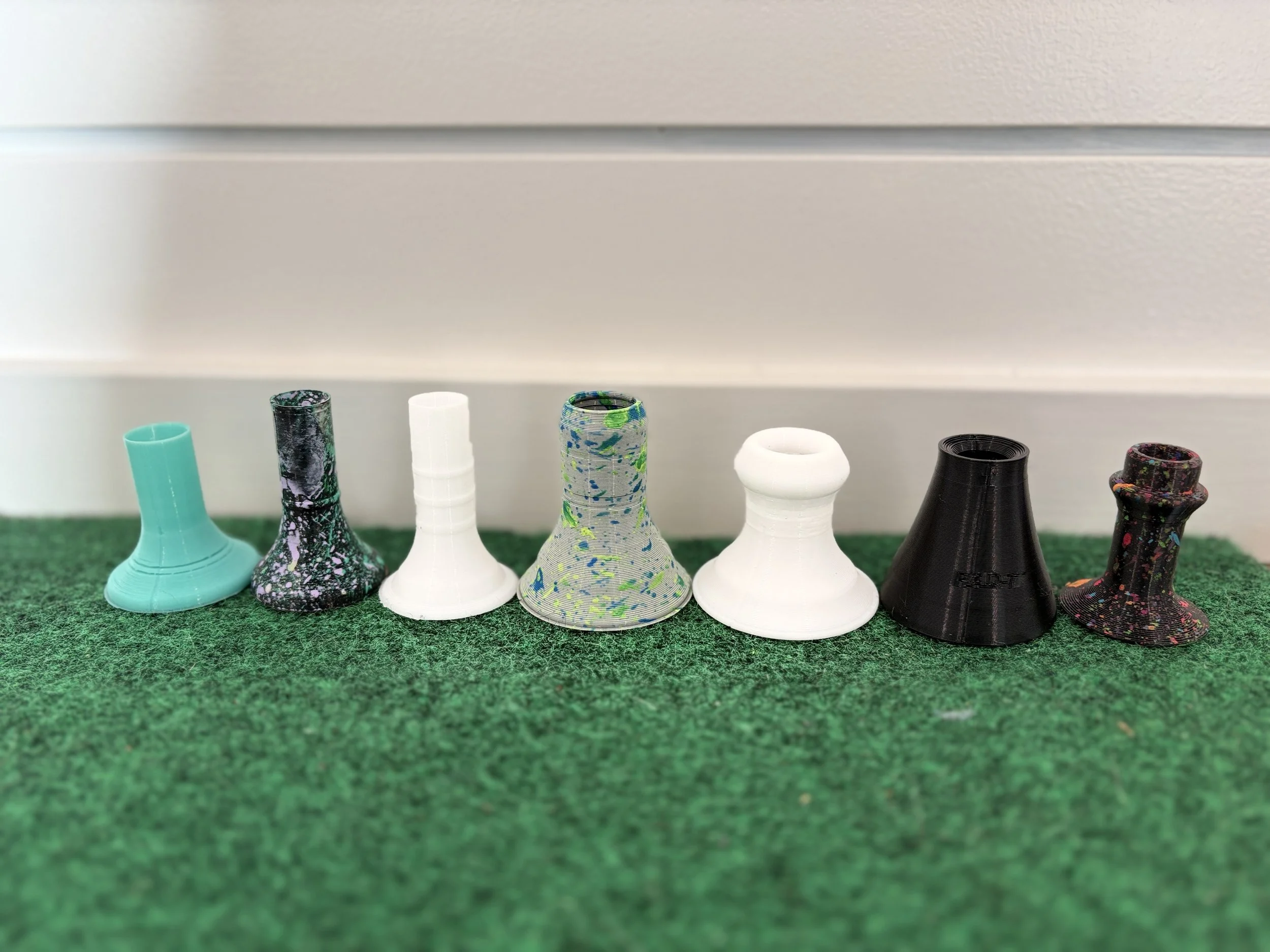 Seven colorful 3D-printed chess pieces lined up on green carpet, against a plain background.