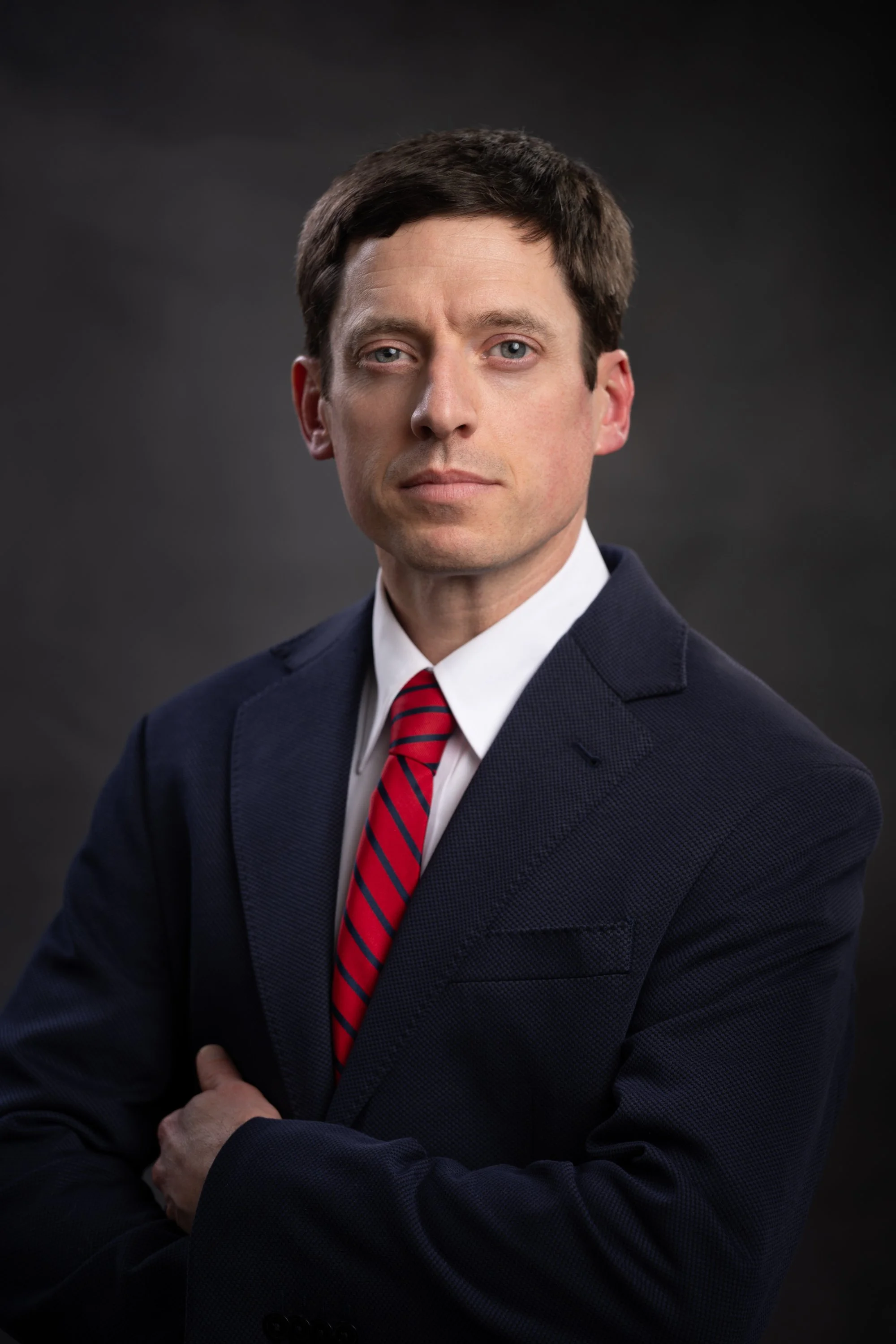 Portrait of a man wearing a navy blue suit, white shirt, and red striped tie against a dark background.