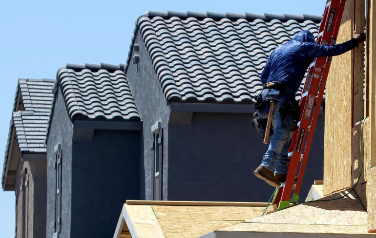 Construction worker wearing a blue jacket on a red ladder working on the side of a house under construction.