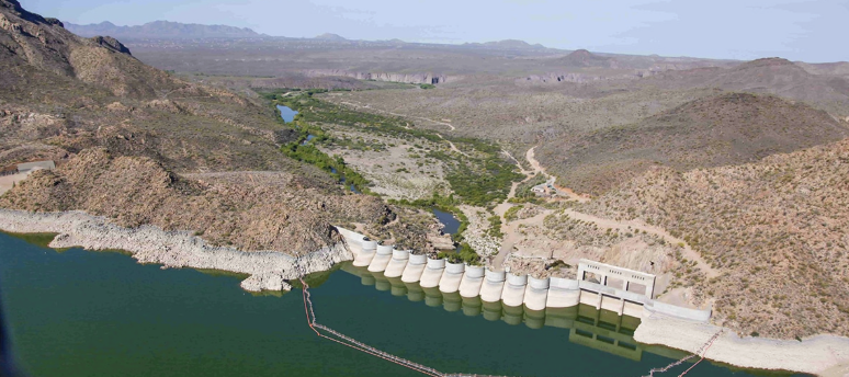 View of a dam with numerous spillway gates holding back water, surrounded by arid, hilly terrain with sparse vegetation.