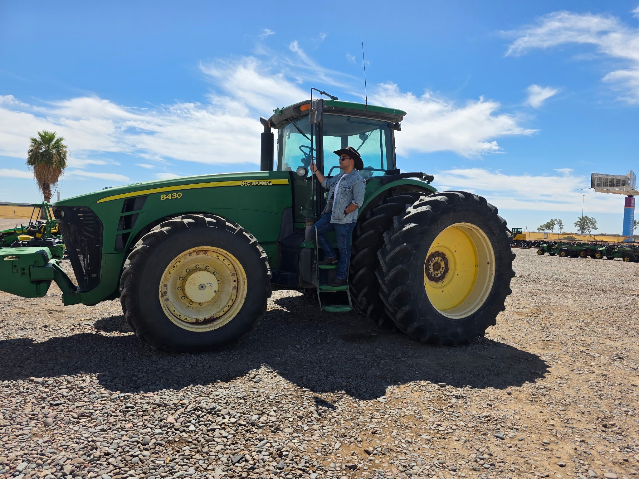 A person standing on the steps of a large green John Deere tractor in a gravel lot on a sunny day, with a palm tree and blue sky with clouds in the background.