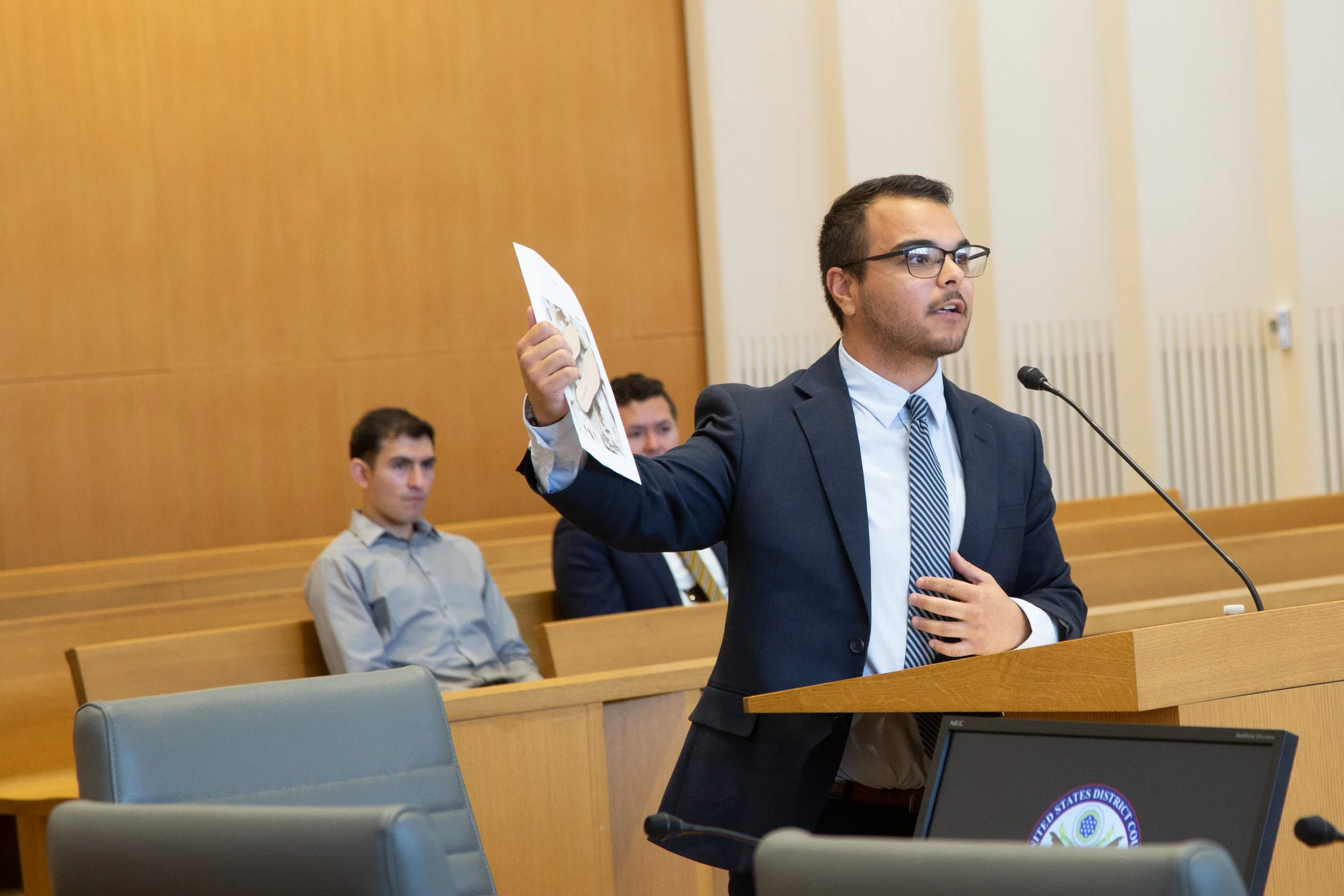 A man in a suit and glasses standing at a podium, holding a printed document, speaking into a microphone in a courtroom with seated individuals in the background.