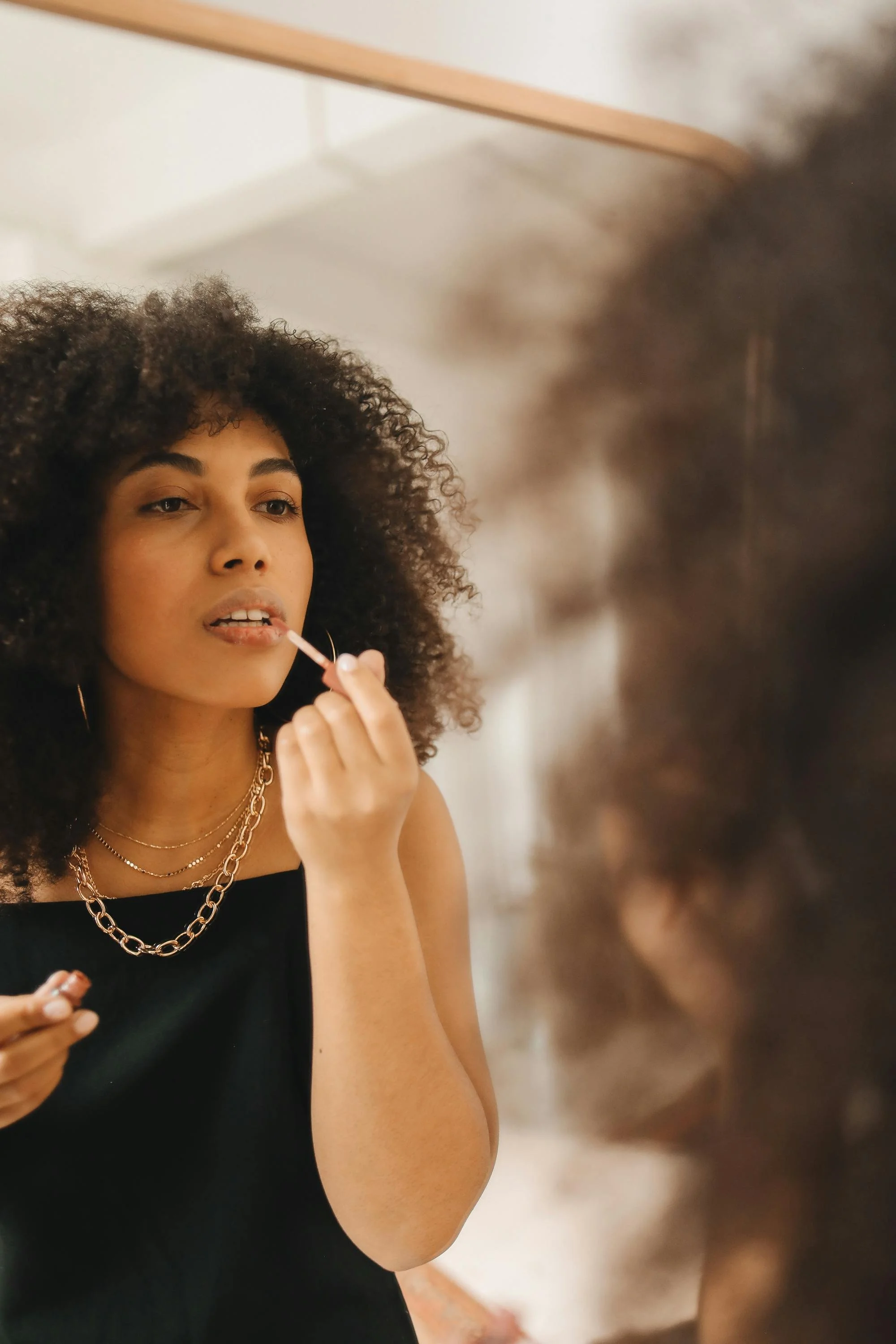 Young Black woman applying lip gloss in a mirror with a distant expression, representing why surface self-care doesn't reach the deeper capacity problem of burnout