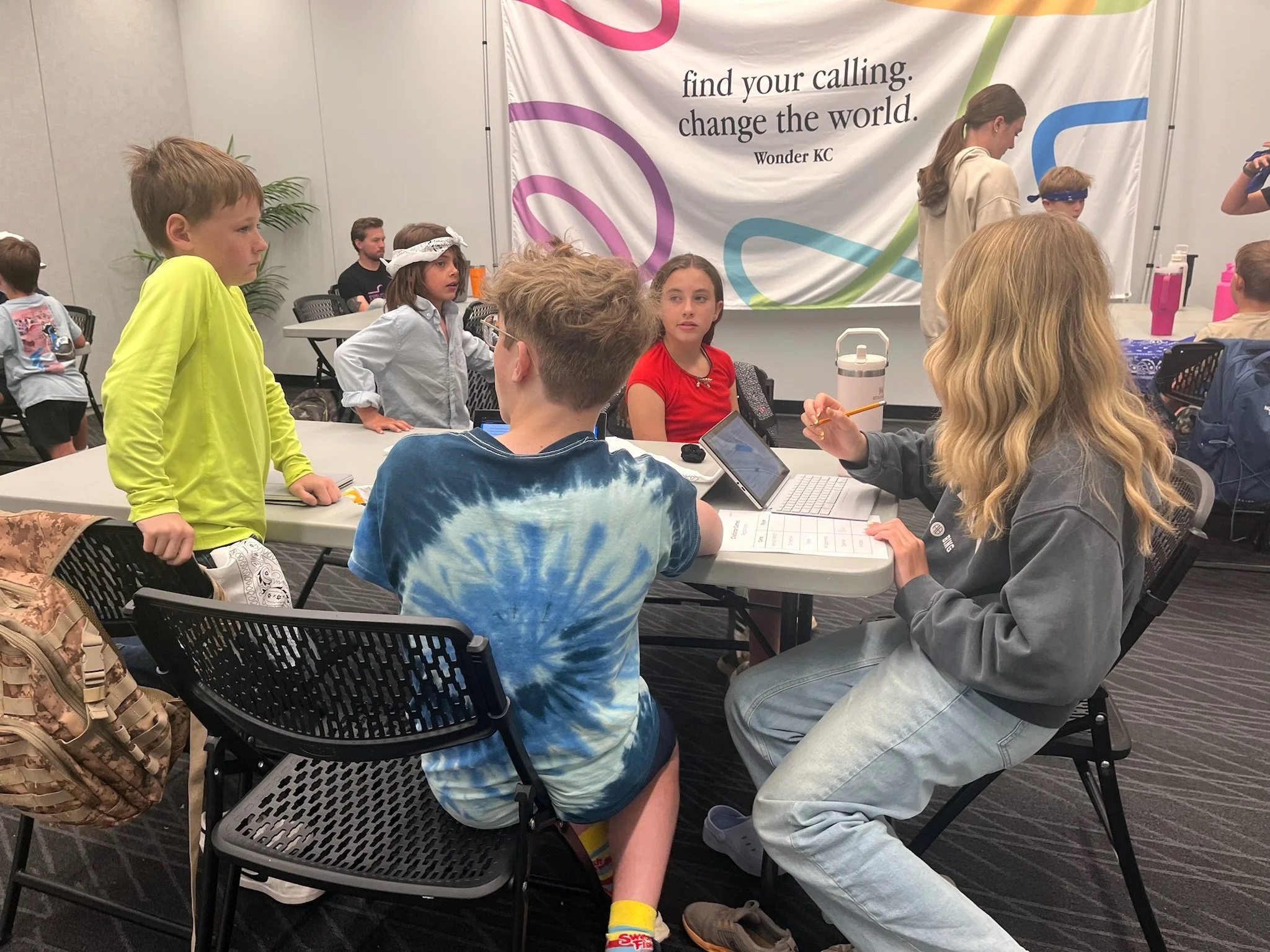 Group of children and a woman in a classroom or workshop setting with a large banner on the wall that reads, 'find your calling. change the world.'