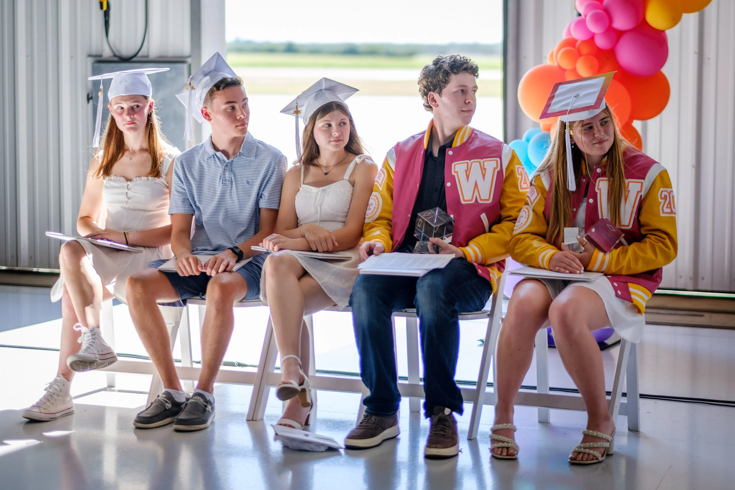 Group of five teenagers sitting on chairs at a graduation ceremony, three wearing white graduation caps and gowns, two dressed in colorful letterman jackets, with balloons in the background.
