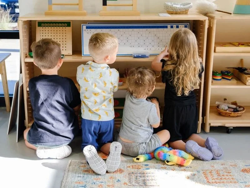Four children sitting on a colorful rug, playing with Montessori materials on a wooden shelf