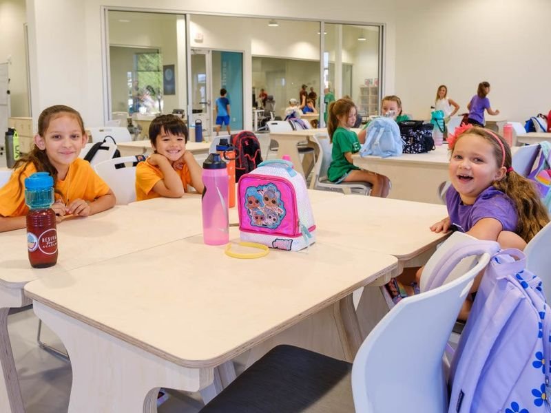 Young children sitting at tables in a bright classroom or cafeteria, smiling and eating, with backpacks and water bottles on the table.