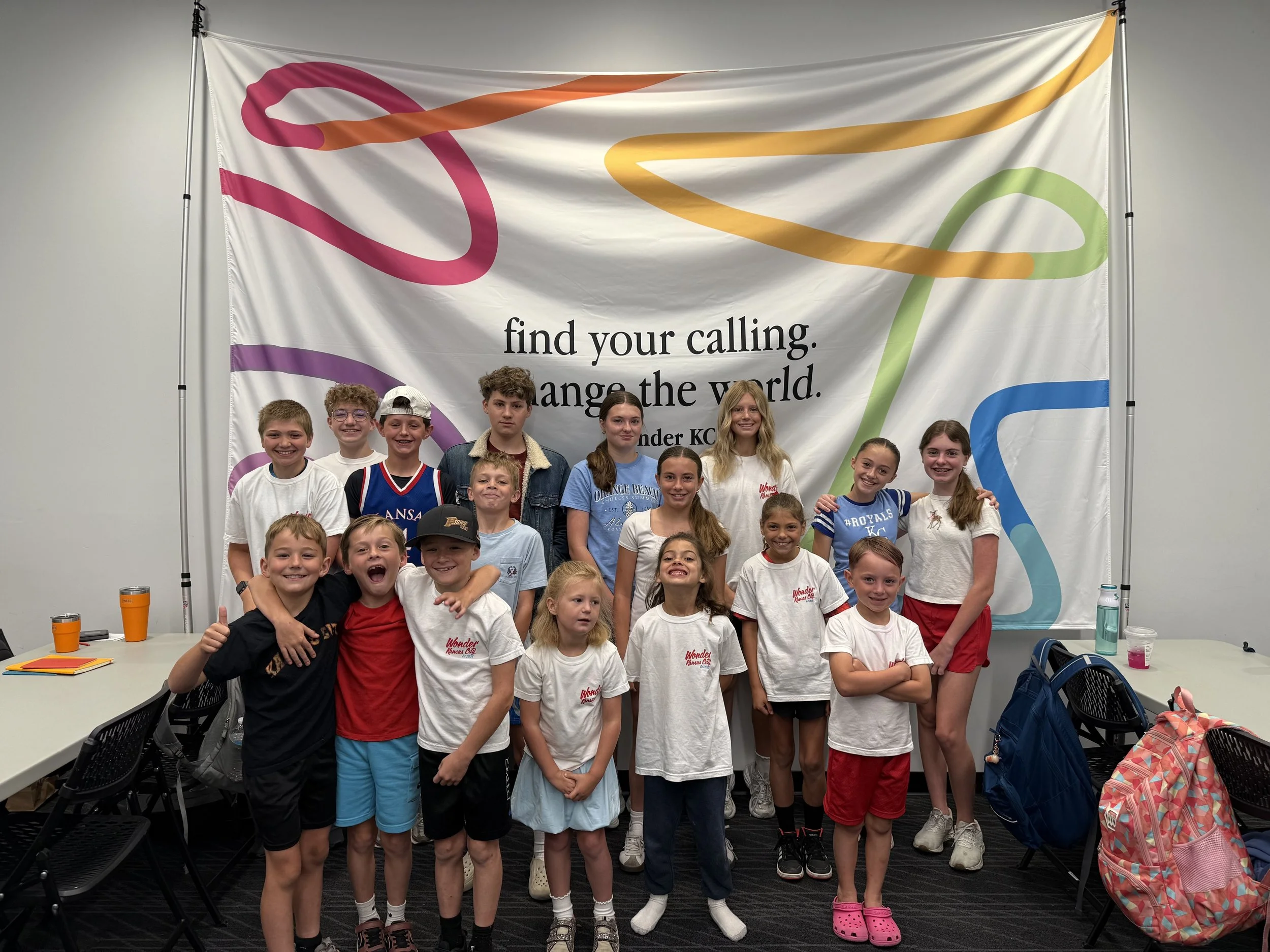 Group of children standing together in front of a banner that reads, "find your calling, change the world." They are smiling and wearing casual clothes, with backpacks and drinks on the table nearby.