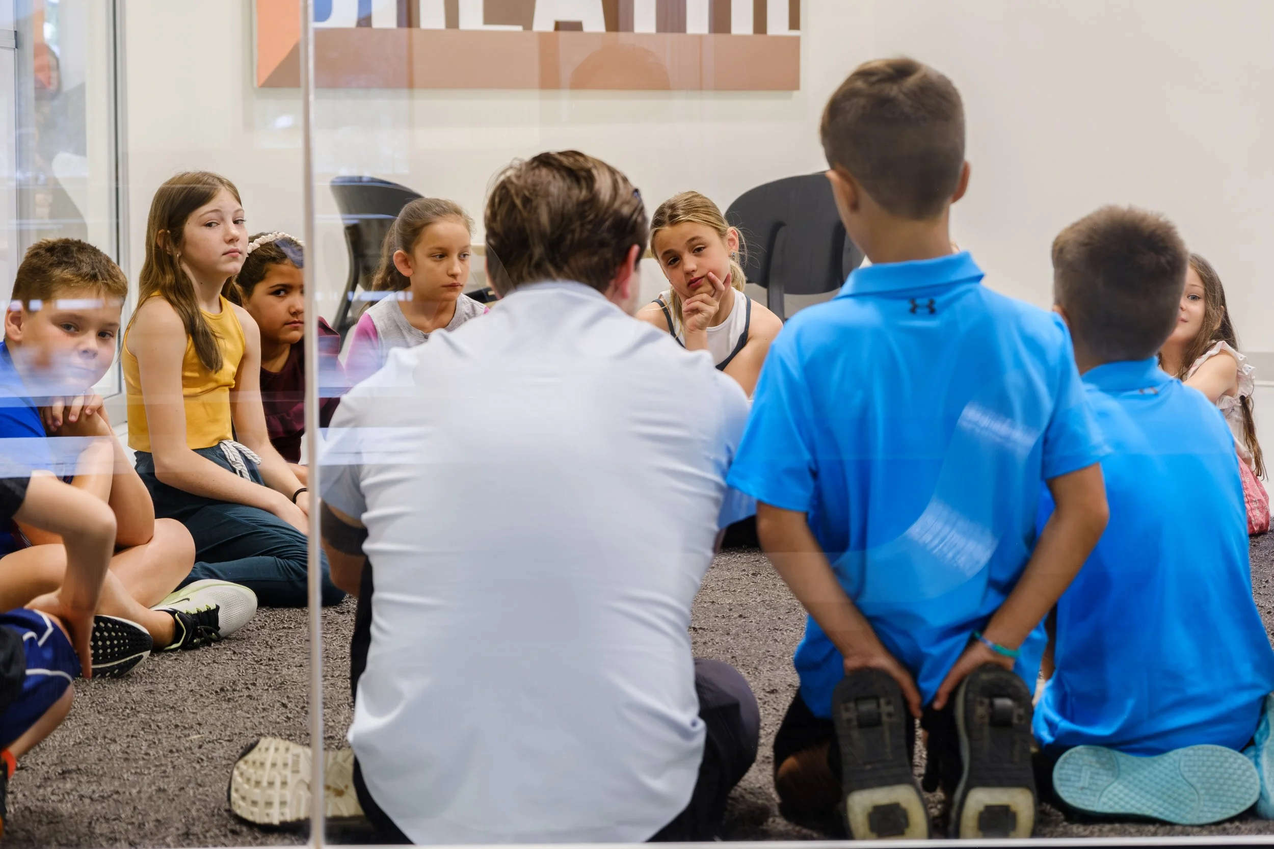 Children sitting on the floor against a glass divider, listening to a man with a beard and short dark hair, wearing a white shirt, who is speaking to them in a classroom setting.