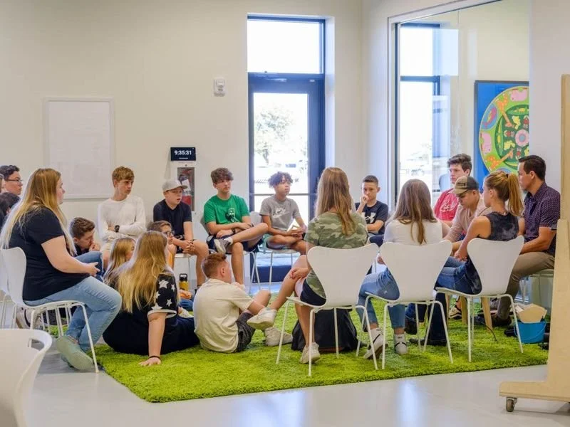 Group of children and teenagers sitting in a circle during a group activity or discussion in a brightly lit room with large windows and colorful artwork.