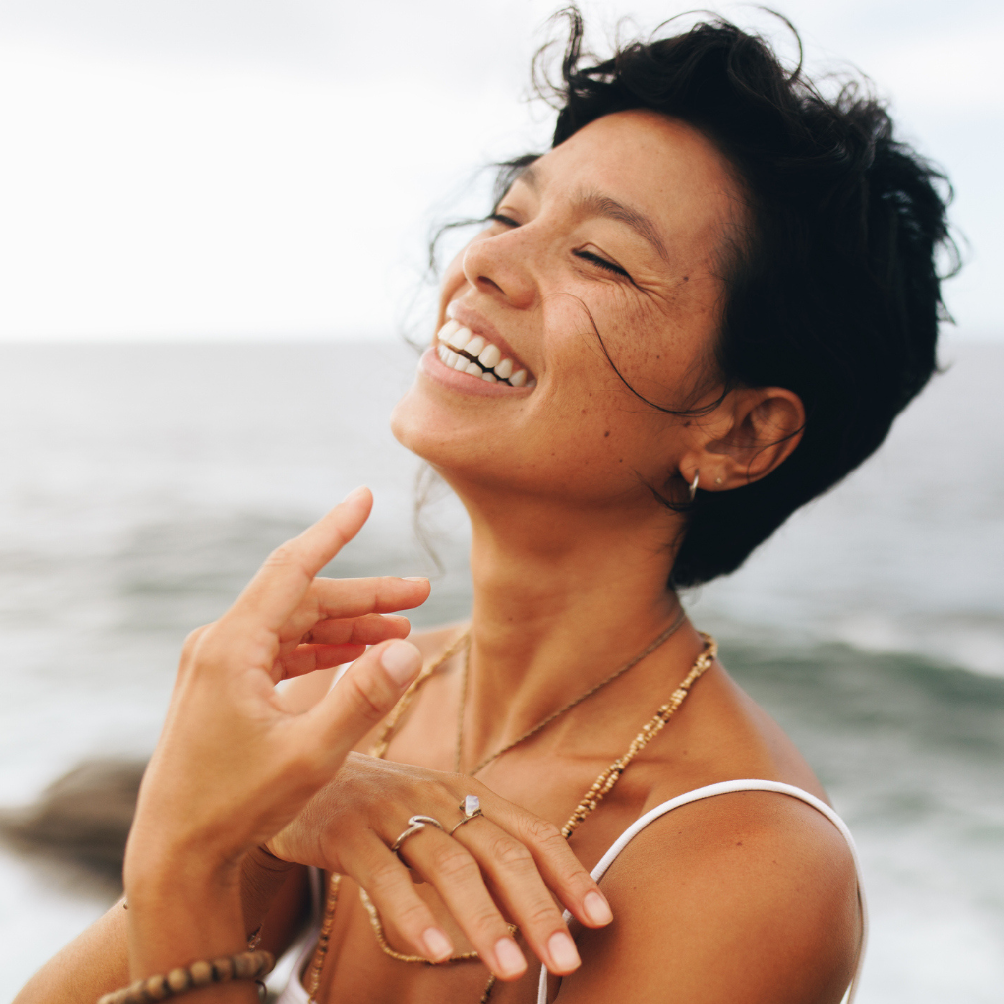 Woman with short dark hair and jewelry smiles outdoors near the ocean, wearing a white top showcasing skincare for mature skin.