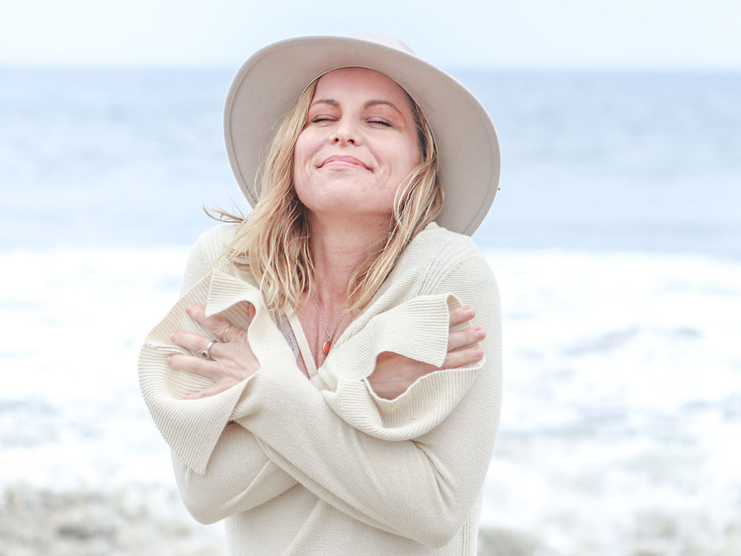 Woman hugging herself at the beach, wearing a wide-brimmed hat and cream-colored sweater, with a smile and closed eyes.