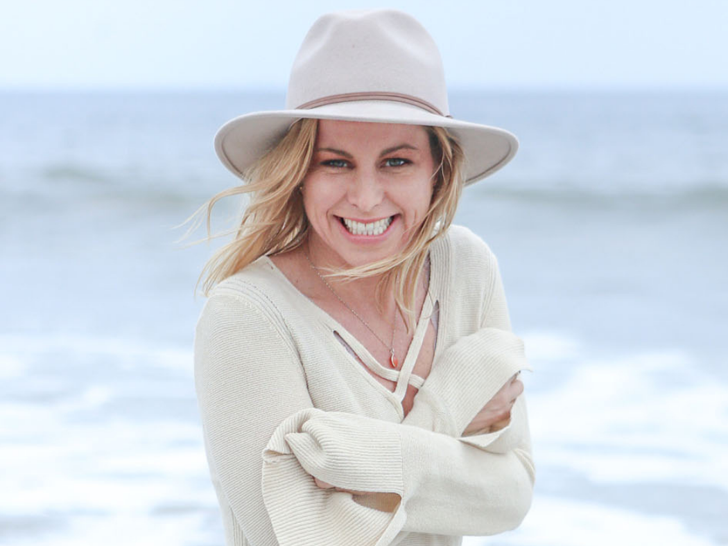 A smiling woman with blonde hair wearing a beige hat and sweater standing on the beach with waves in the background.