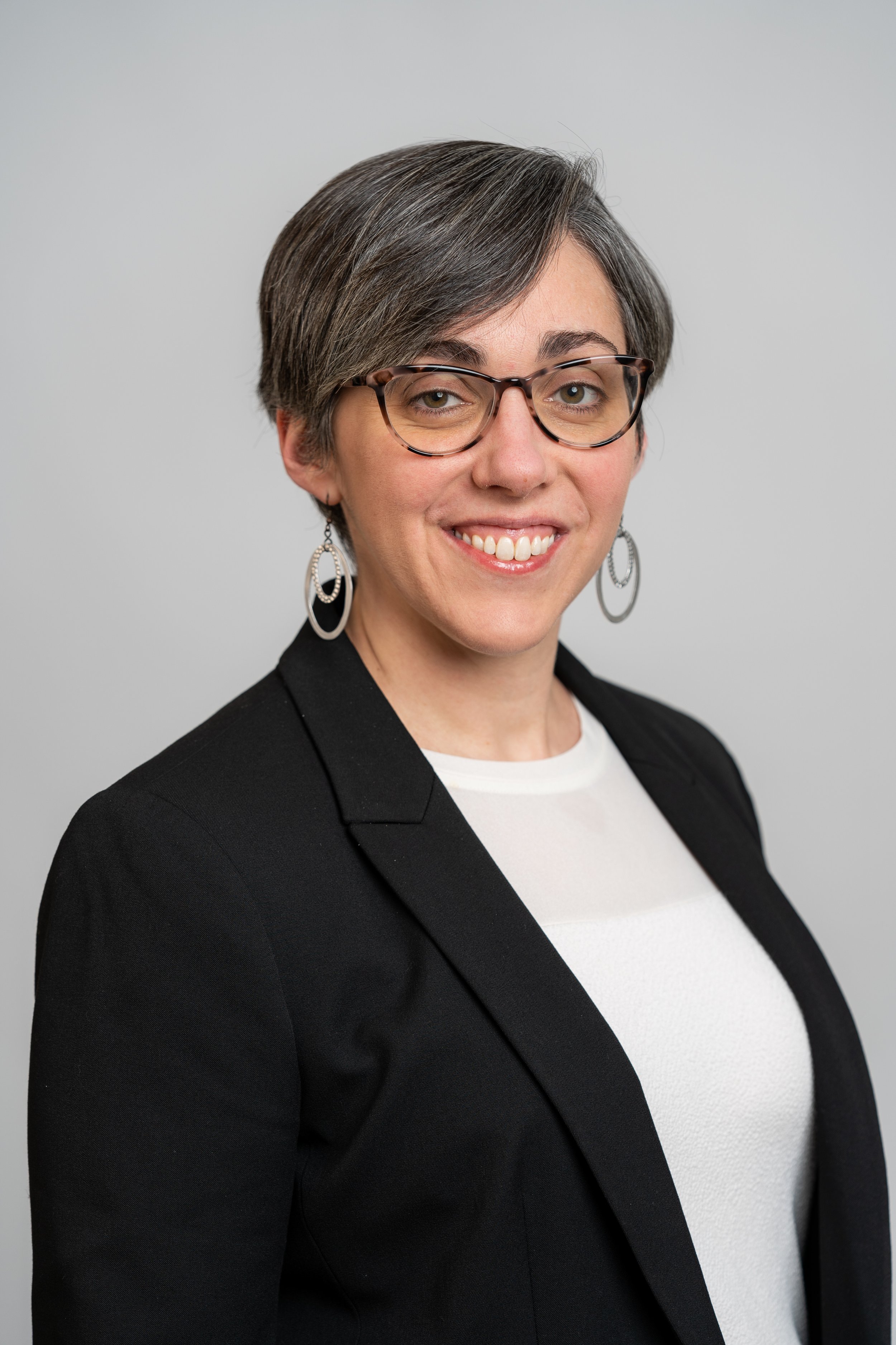 Headshot of Christine Gavin Teahan, a woman with short brown and gray hair, glasses, and hoop earrings, wearing a black blazer over a white top, smiling at the camera against a plain gray background.