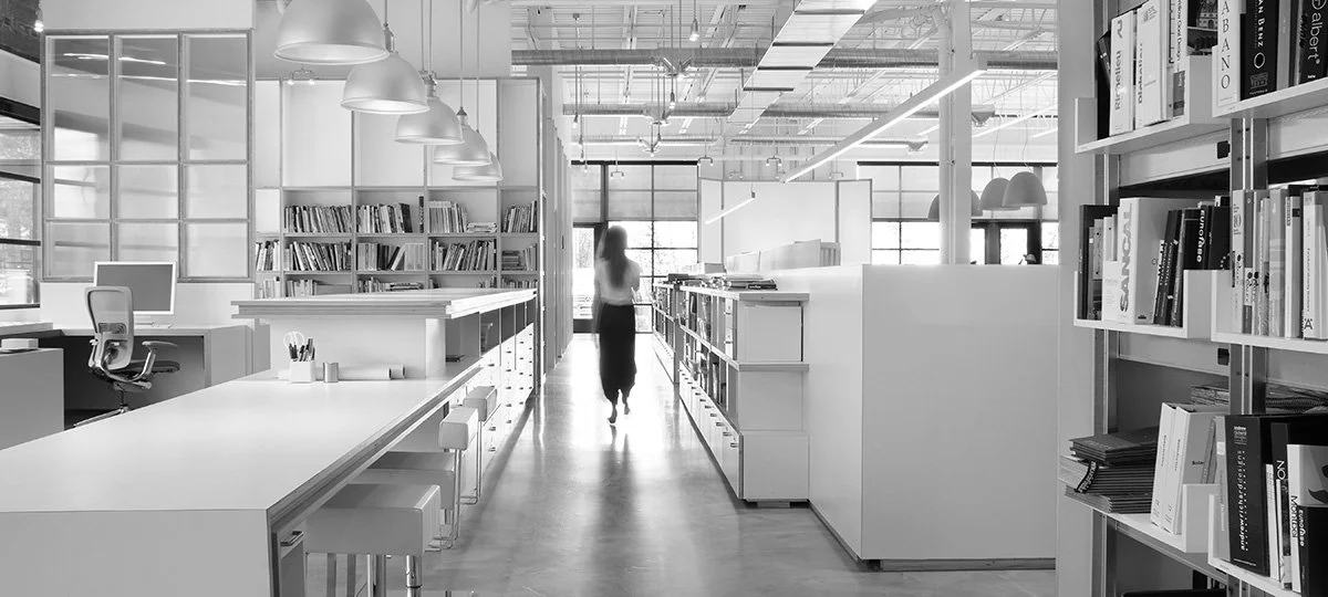 A modern, well-lit library or office space with shelves of books, a white table with chairs, and a person walking down the center aisle.