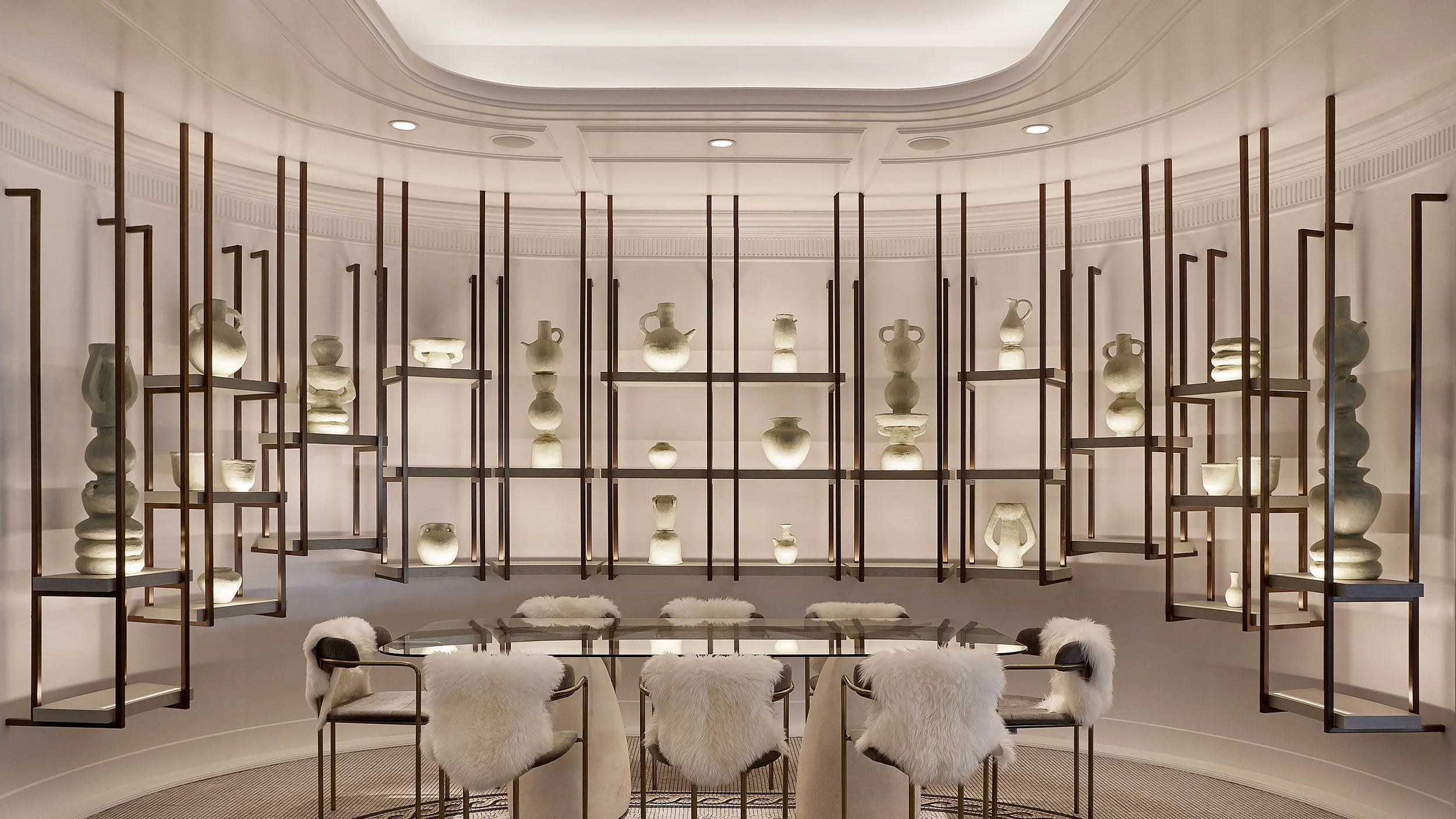 A contemporary room displaying a collection of ancient pottery and vases on black geometric metal shelves, with four white fluffy chairs around a round glass table in the foreground.