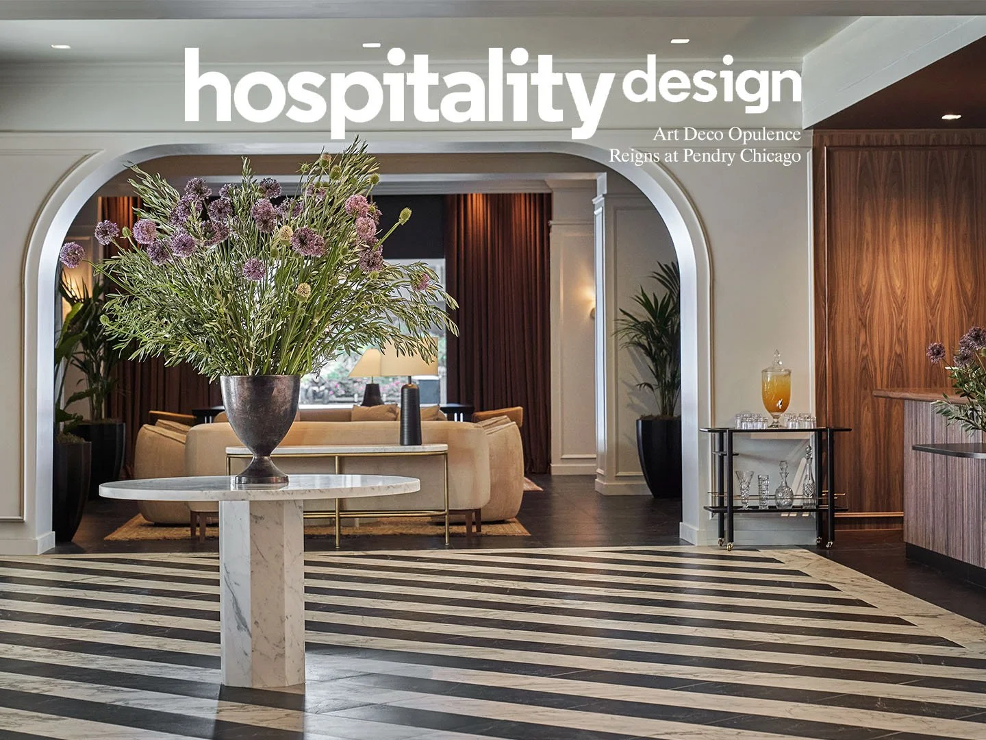 Elegant hotel lobby with a large flower arrangement on a marble table, a sitting area with beige sofas, and a bar area with drinks in the background.