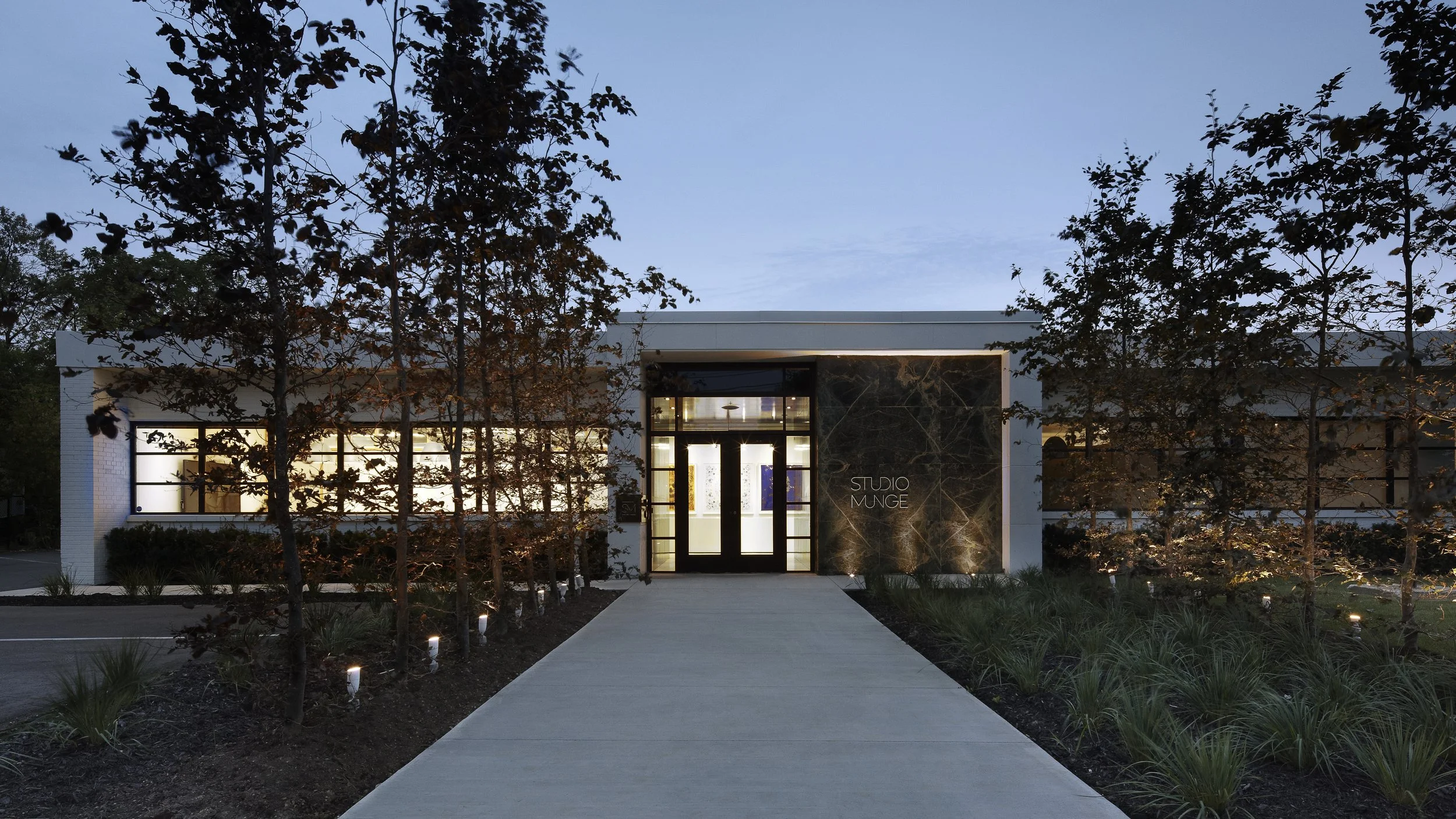 Exterior of a modern building with large windows, trees in front, and a sign that reads B Studio Munge, illuminated landscaping lights along a paved walkway at dusk.