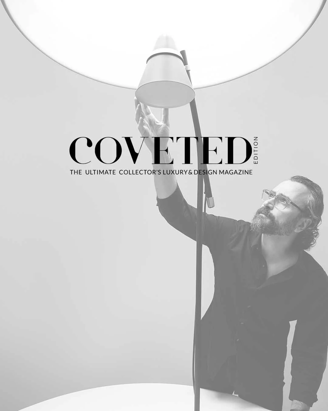 Black and white magazine cover featuring a man with glasses and a beard adjusting a large lamp above his head. The text on the cover reads 'COVETED' with a subtitle 'The ultimate collector's luxury & design magazine.'