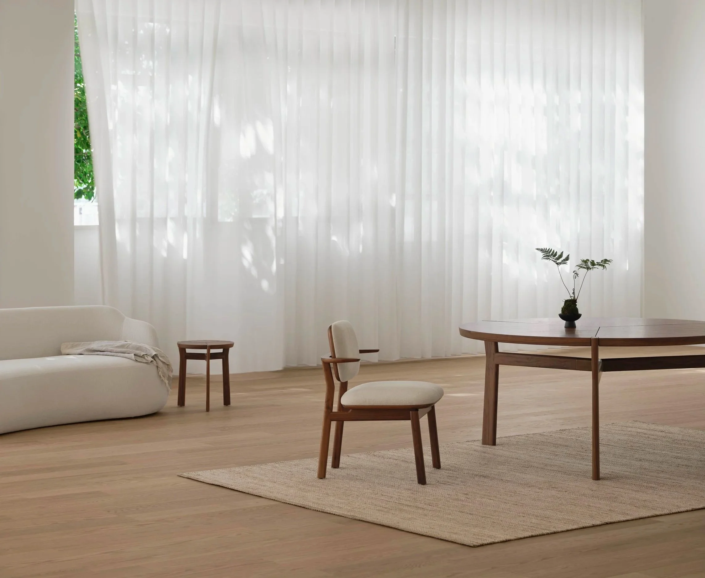 A room with a large window covered with white sheer curtains, a white sofa with a blanket, wooden chairs, a round wooden table with a potted plant, and a beige area rug on light wooden floor.