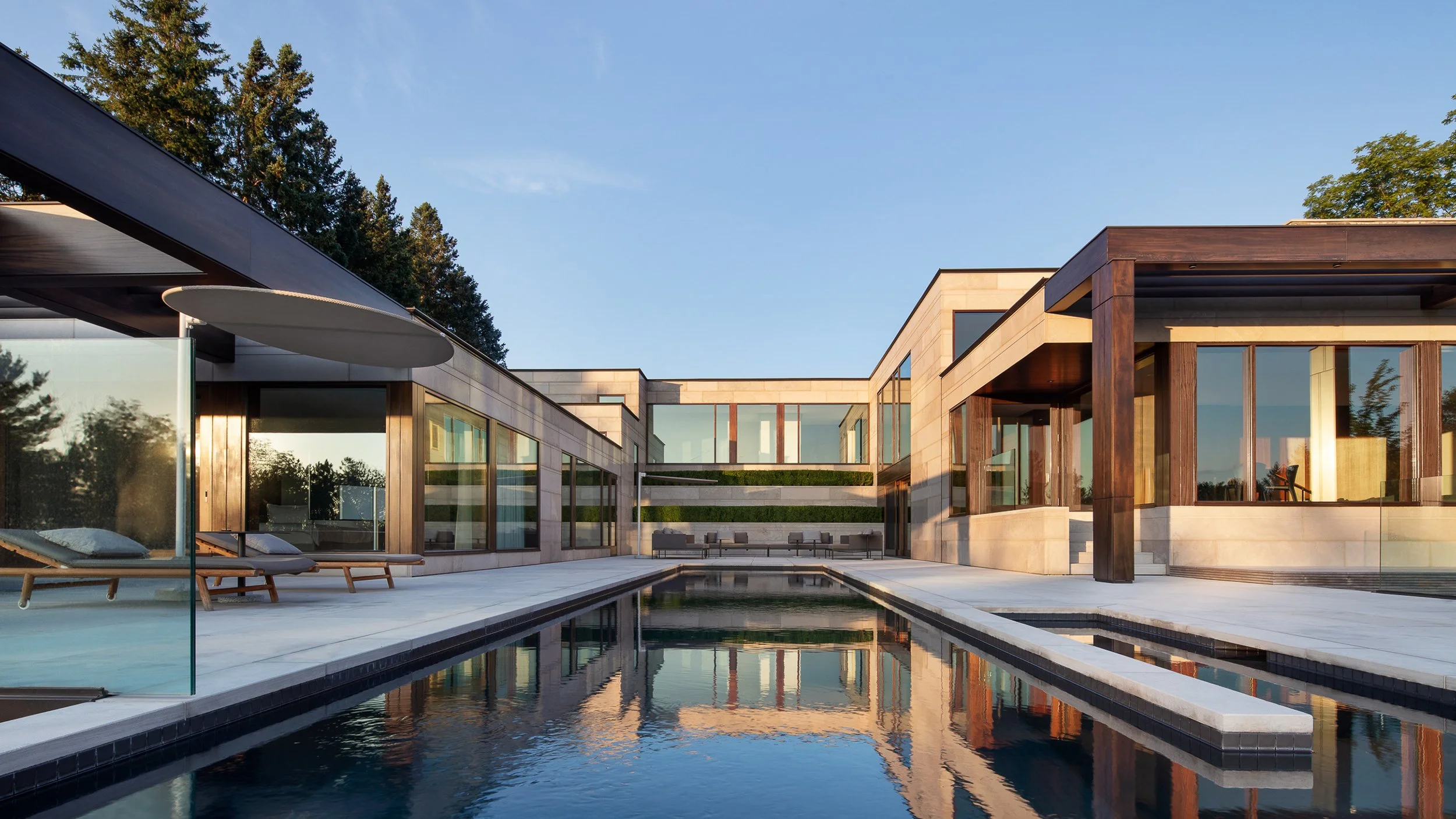 Modern luxury house with large glass windows, featuring a pool in the foreground and greenery around, under a clear blue sky.