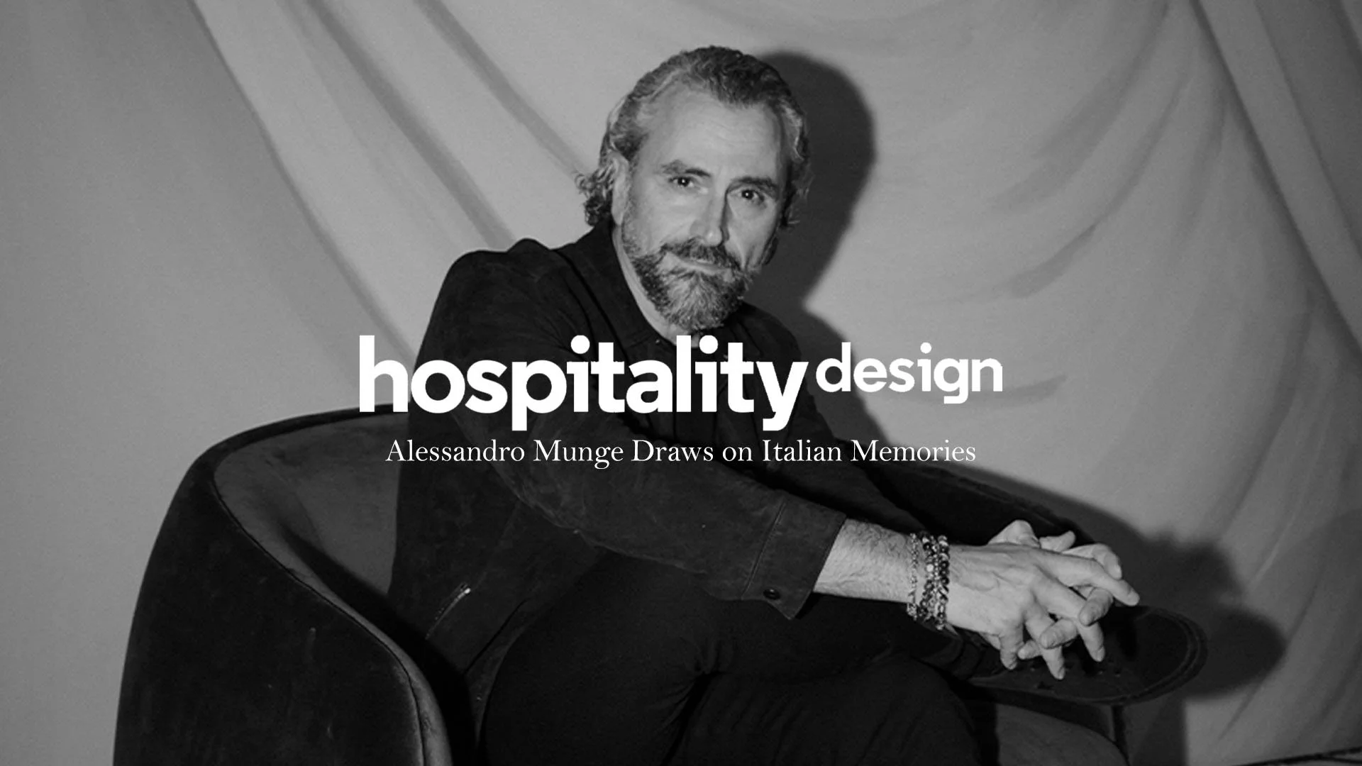 A black and white photo of a man with gray hair and beard sitting at a piano, looking at the camera, with text overlay that reads 'hospitality design' and 'Alessandro Munge Draws on Italian Memories.'