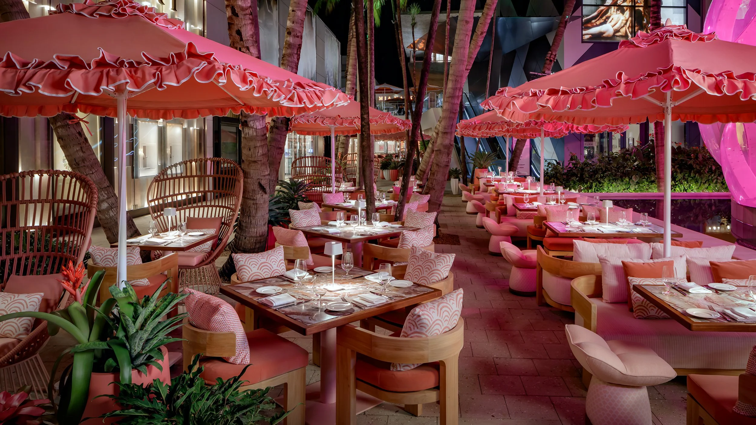 Outdoor restaurant patio with pink umbrellas, pink seating, and palm trees, set for dining with tables, chairs, and glassware at night.