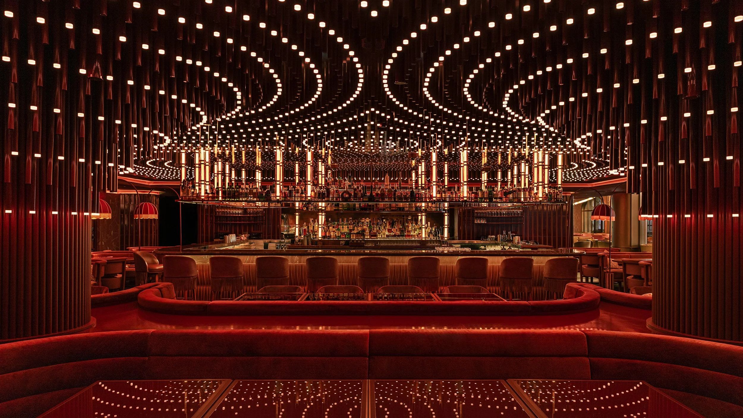 A luxurious bar with a glowing, geometric ceiling design and red velvet seating.