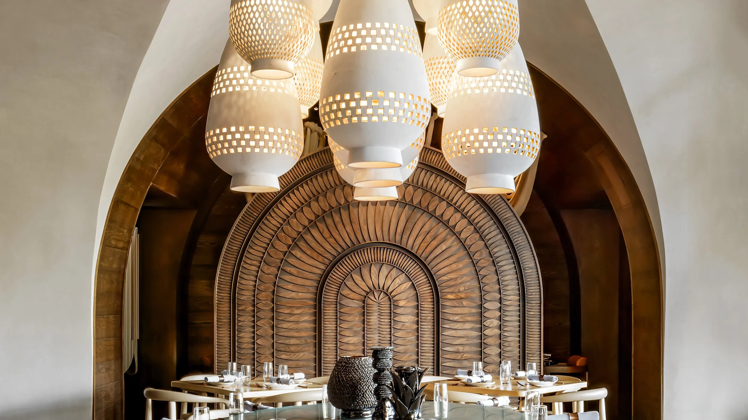 A luxurious dining room with a large wooden table set with glassware and dishes. Above the table hangs a cluster of white, perforated pendant lights. The background features a decorative wooden panel with a circular, sunburst pattern arching over the space, framed by white walls.