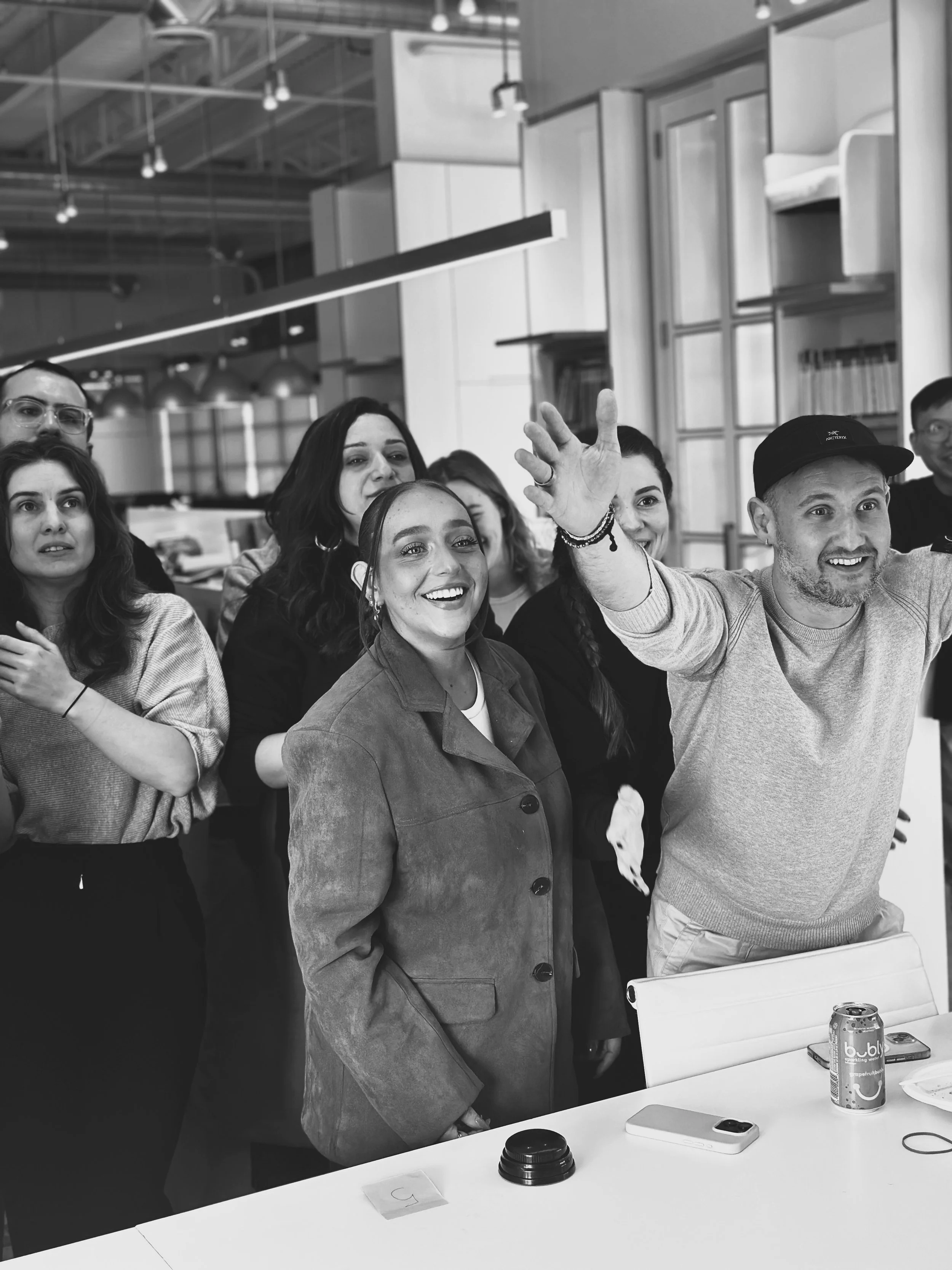 Group of people gathered around a white table in an office, smiling and interacting, with two cans and electronic devices on the table.