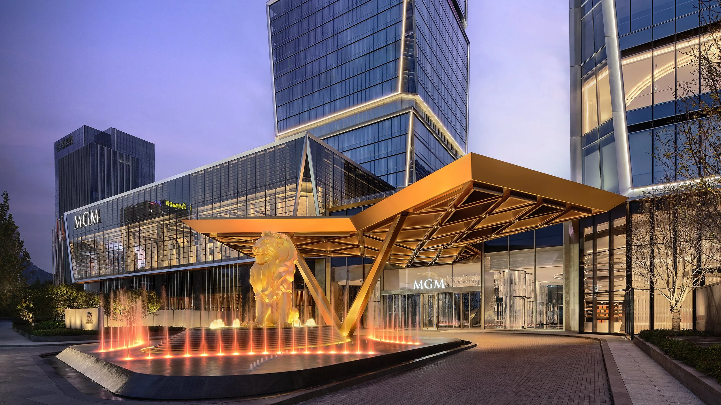 Night view of the exterior of the MGM hotel and casino with a modern glass facade, a decorative lion statue in a fountain, and a canopy entrance.