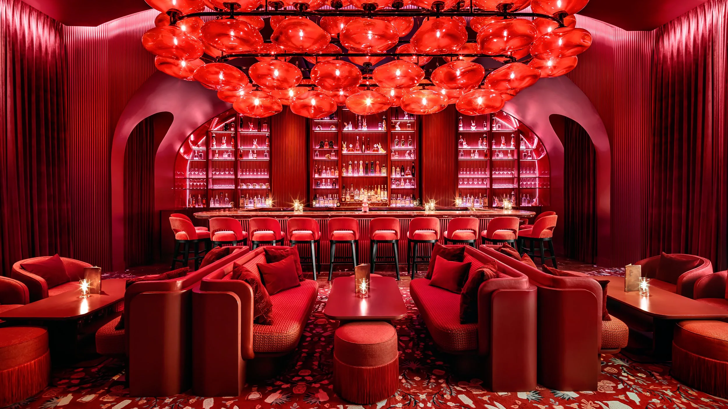 A luxurious bar interior with red lighting, featuring a chandelier of red glass globes, red velvet seating, and a well-stocked bar with bottles displayed on backlit shelves.