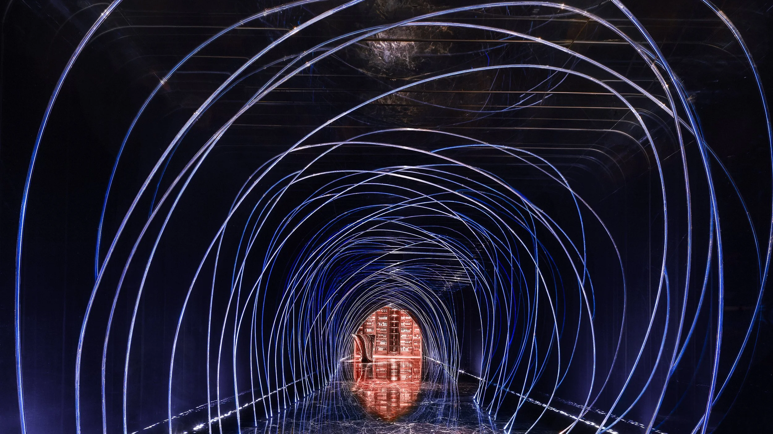 A tunnel with glowing blue and white neon lights forming arches, with a red-lit structure at the end and a reflection on the shiny floor.