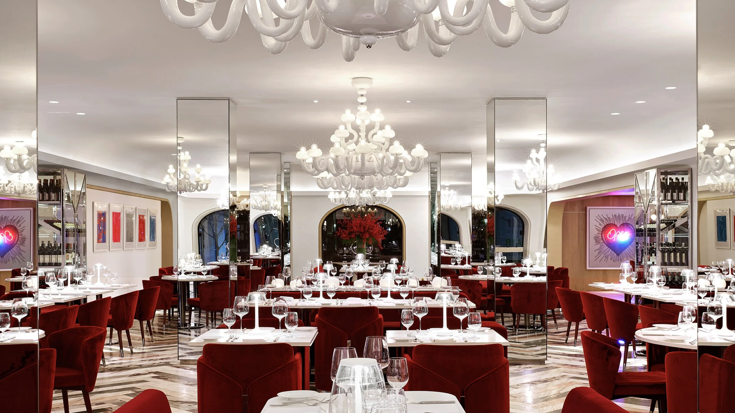 Elegant restaurant interior with red velvet chairs, white tablecloths, decorated chandeliers, mirrored columns, and neon signs, featuring a large red floral centerpiece.