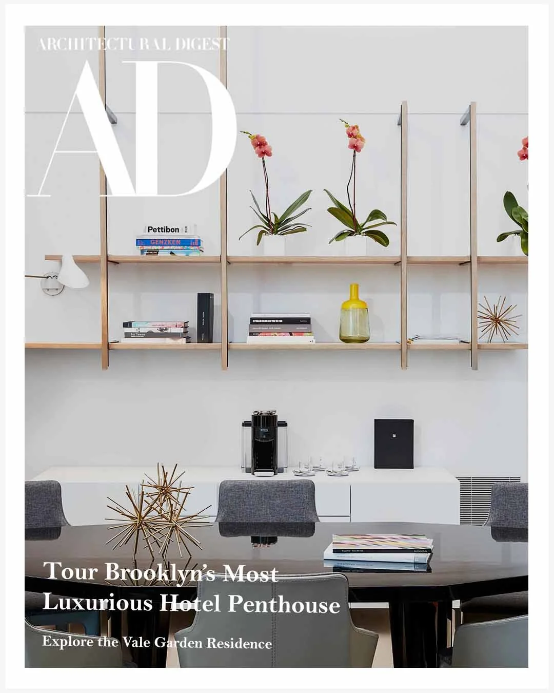 Interior of a modern apartment with a dining table, grey chairs, black and white books, decorative objects, and a wall-mounted shelving unit with potted plants and books.