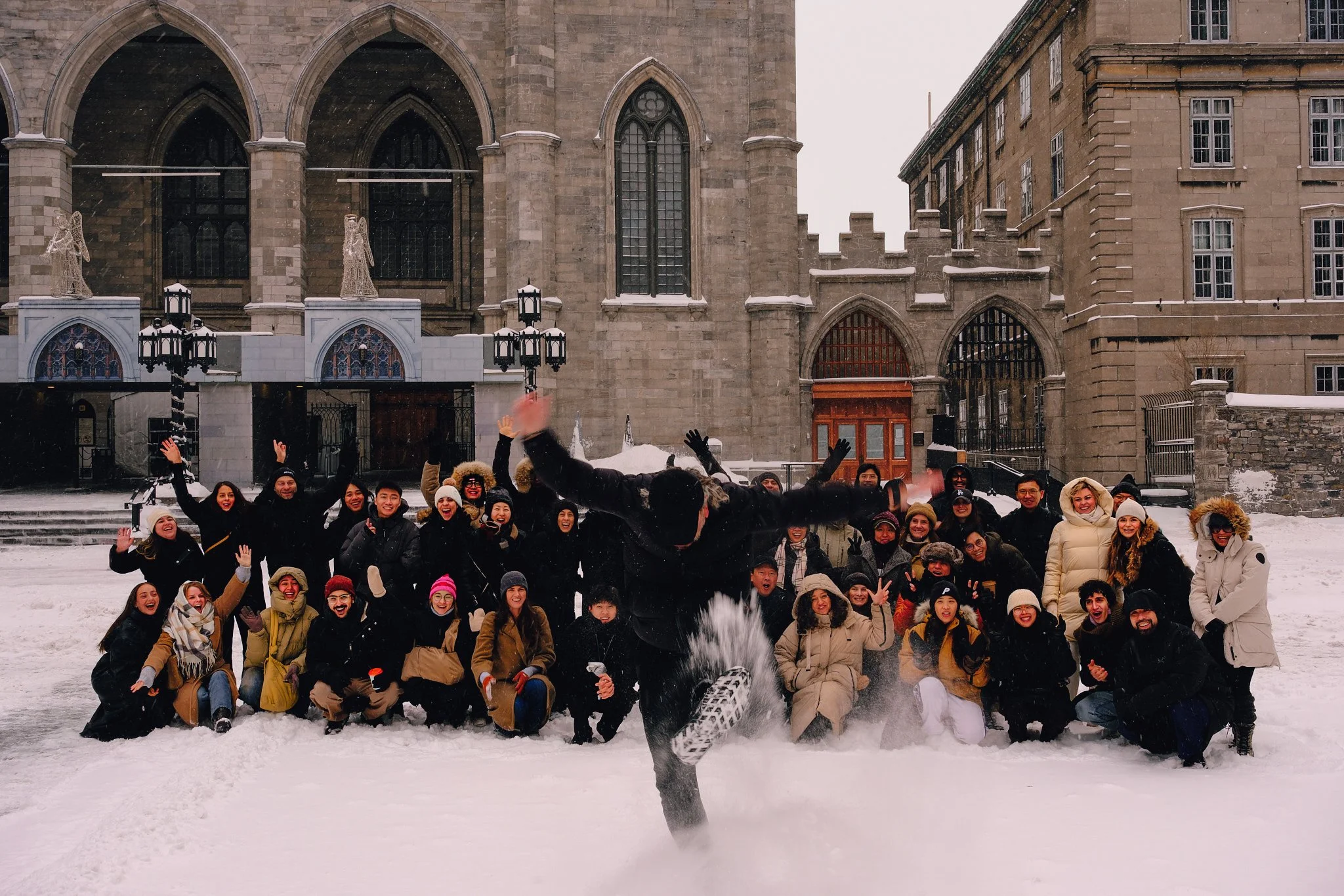 A group of people dressed in winter clothing posing in snow in front of a large historic building with gothic windows and archways, some snow-covered angel decorations hanging outside.