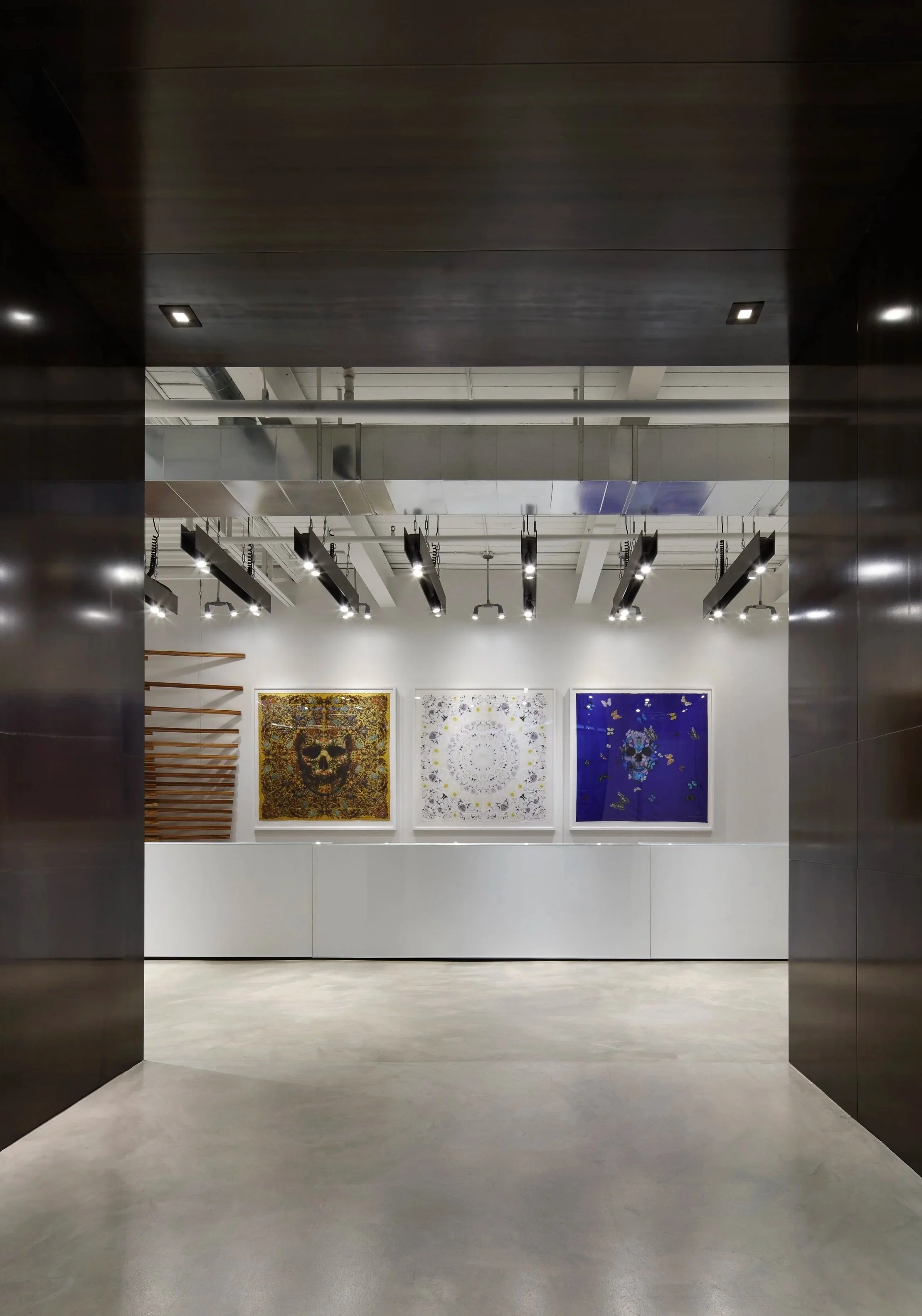 View of an art gallery with three large colorful artworks hanging on the wall, illuminated by ceiling spotlights, and viewed through an open doorway with dark metal framing.