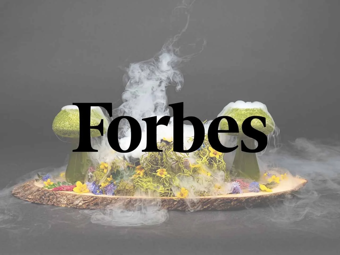 A floating dessert presentation with two green dome-shaped components topped with white foam, surrounded by flowers, greenery, and mist or smoke on a wooden slab.