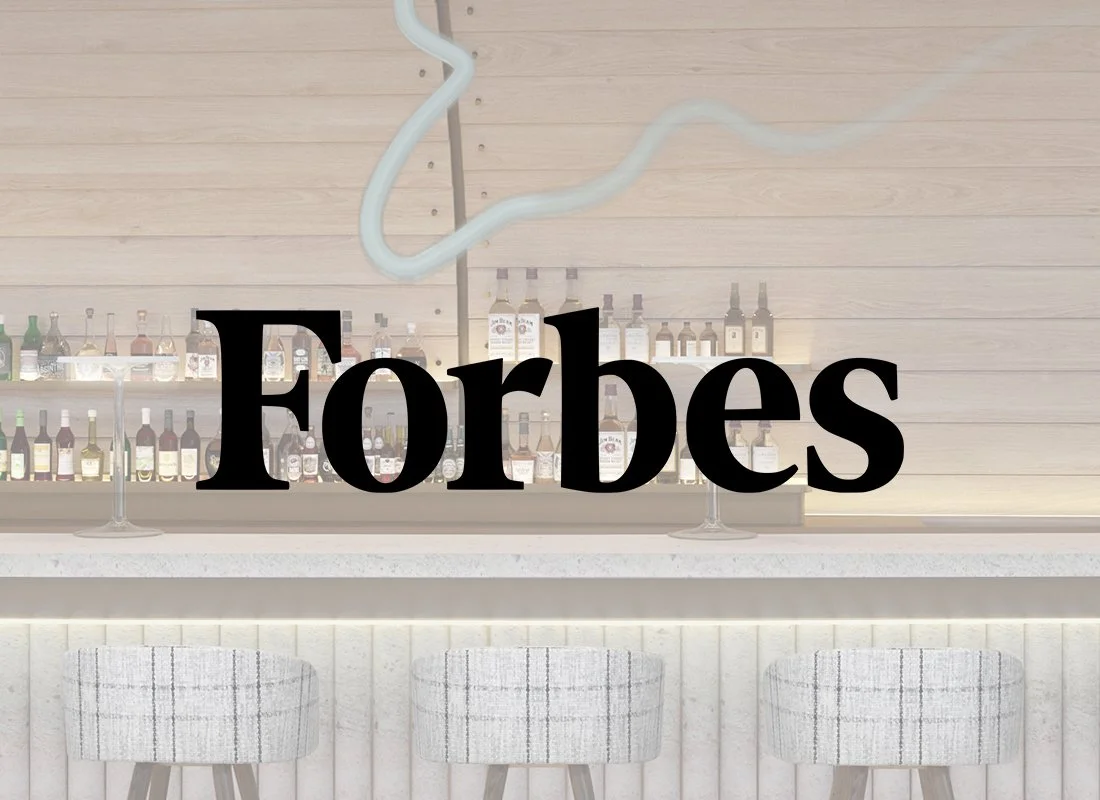 Modern bar counter with three cushioned barstools in front, alcohol bottles on shelves behind, and a decorative neon light shaped like an abstract cloud above. The word 'Forbes' is displayed prominently in the center.
