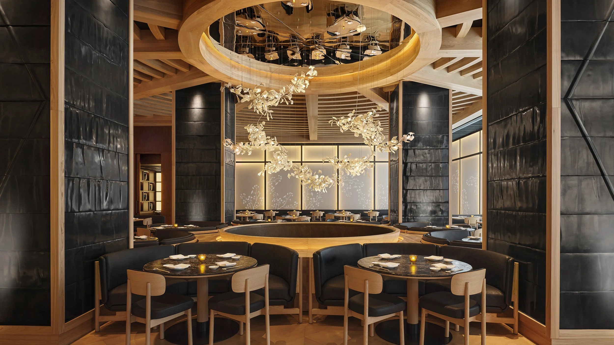 A modern restaurant interior with black circular tables, wooden chairs, decorative lighting, and dark walls, featuring a large round chandelier with white floral accents and a reflective ceiling with black and white elements.