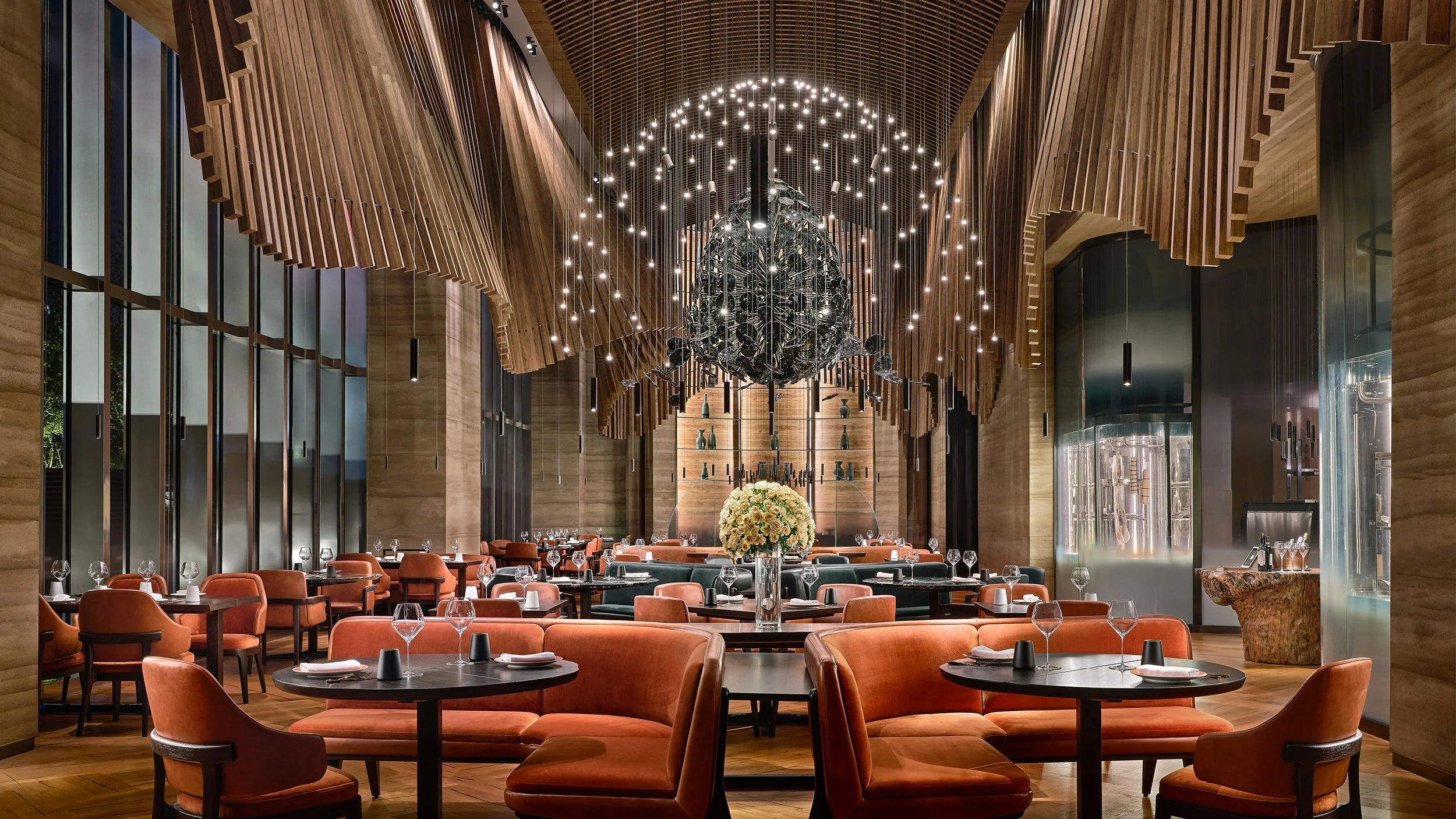 Elegant restaurant interior with warm wooden walls, modern hanging light fixtures, and round tables set with glassware and napkins.