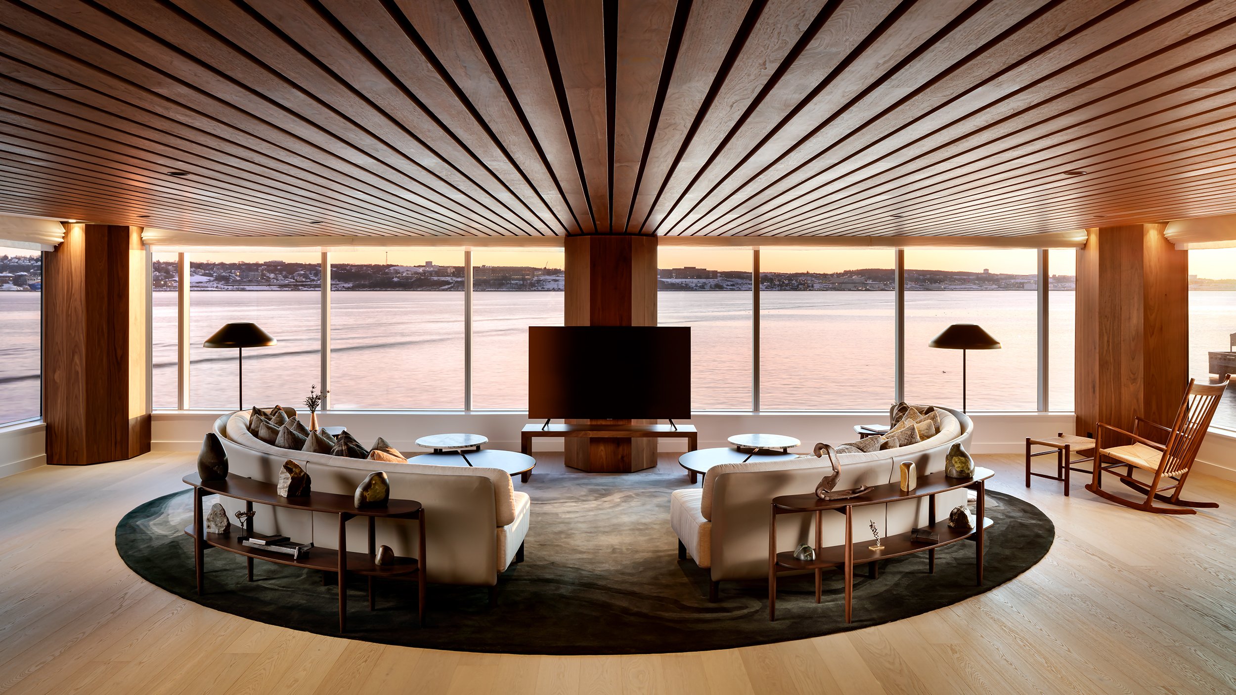 Modern living room with large windows overlooking a body of water at sunset, featuring a circular sofa, armchairs, coffee tables, lamps, and decorative items.
