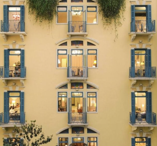 Front view of a yellow building with multiple blue shuttered windows and small balconies, some with glass doors or open windows, illuminated interior rooms, and green plants hanging from the top.