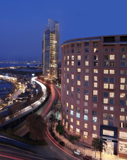 Nighttime view of modern high-rise buildings and busy city streets with light trails from moving vehicles in an urban area.