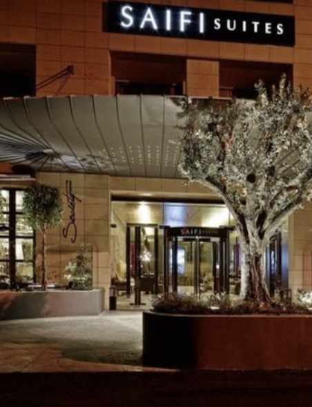 Nighttime view of the entrance to SAIF Suites hotel with a large tree in a planter in front, a curved canopy over the entrance, and illuminated signage.